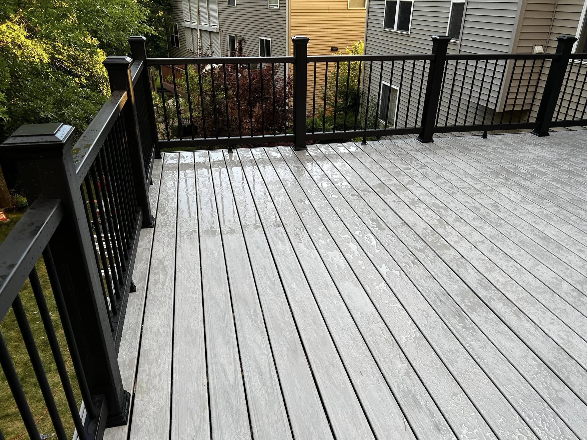 A wooden deck with wet planks and a black metal railing, overlooking trees and neighboring buildings.