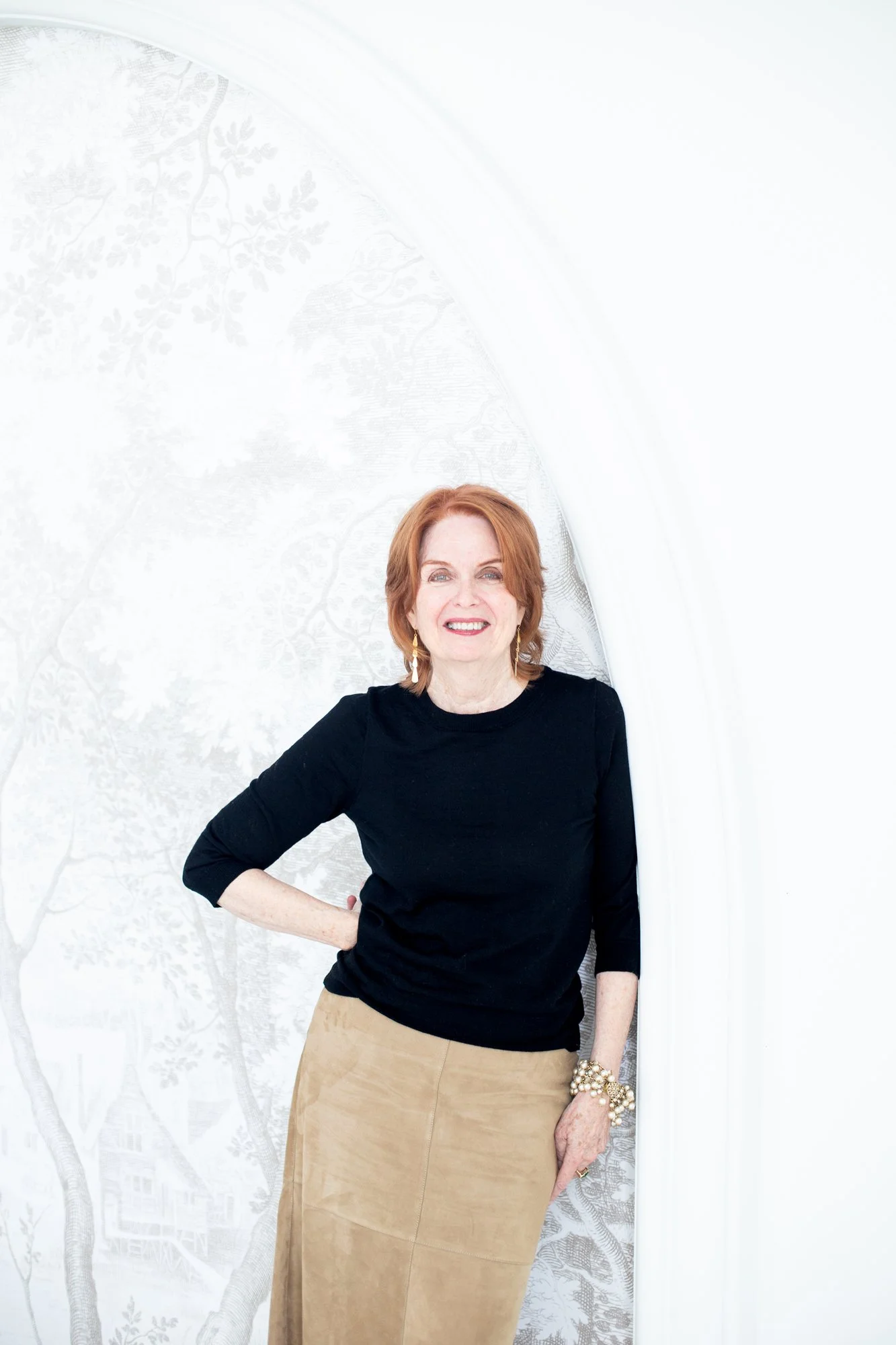 A woman with red hair, wearing a black top and beige skirt, standing in front of a white wall with a tree design, smiling at the camera.
