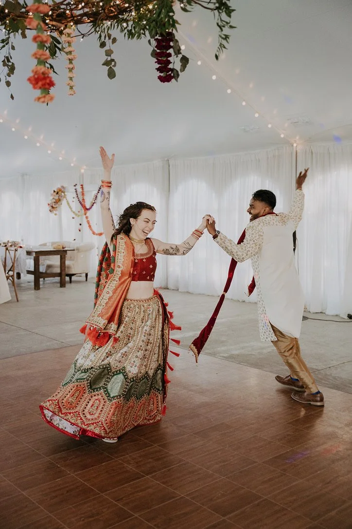 A couple dancing at a wedding reception inside a decorated event tent. The woman is wearing traditional colorful Indian attire, while the man is dressed in a cream-colored kurta with a maroon dupatta. They are smiling and holding hands while dancing.