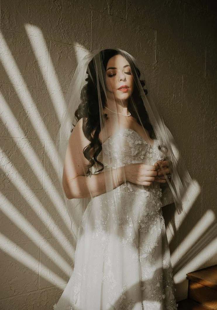 A bride with long dark hair and a wedding dress with lace details, standing against a wall with sunlight creating shadow patterns.