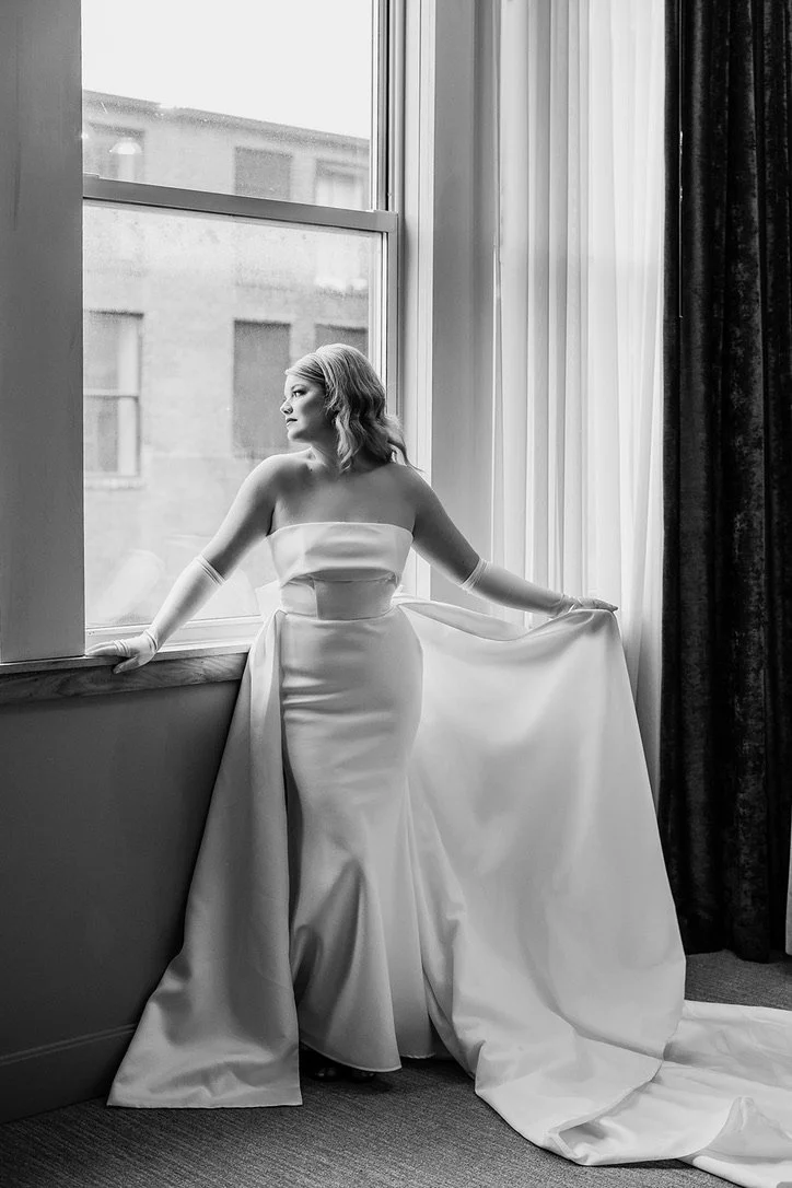 A woman in a strapless wedding dress standing by a large window, looking outside, with her hand on the window sill and the other holding the train of her dress.