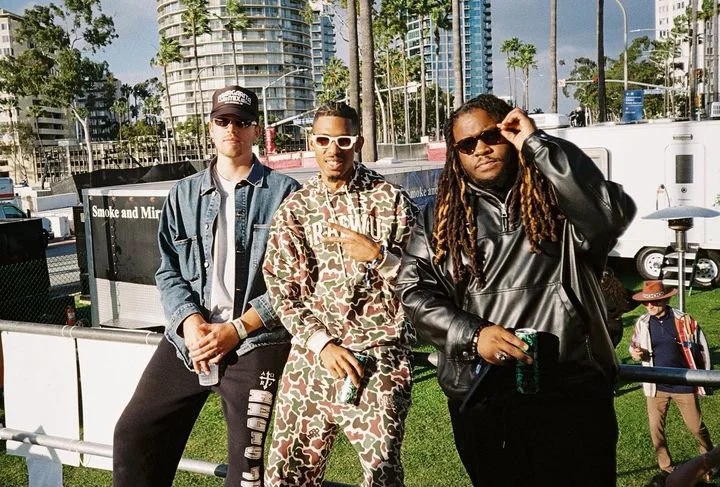 Three people standing outdoors in an urban park with high-rise buildings and palm trees in the background. They are wearing casual and stylish clothing, with two wearing sunglasses. One is holding a drink, and another is showing a hand gesture.