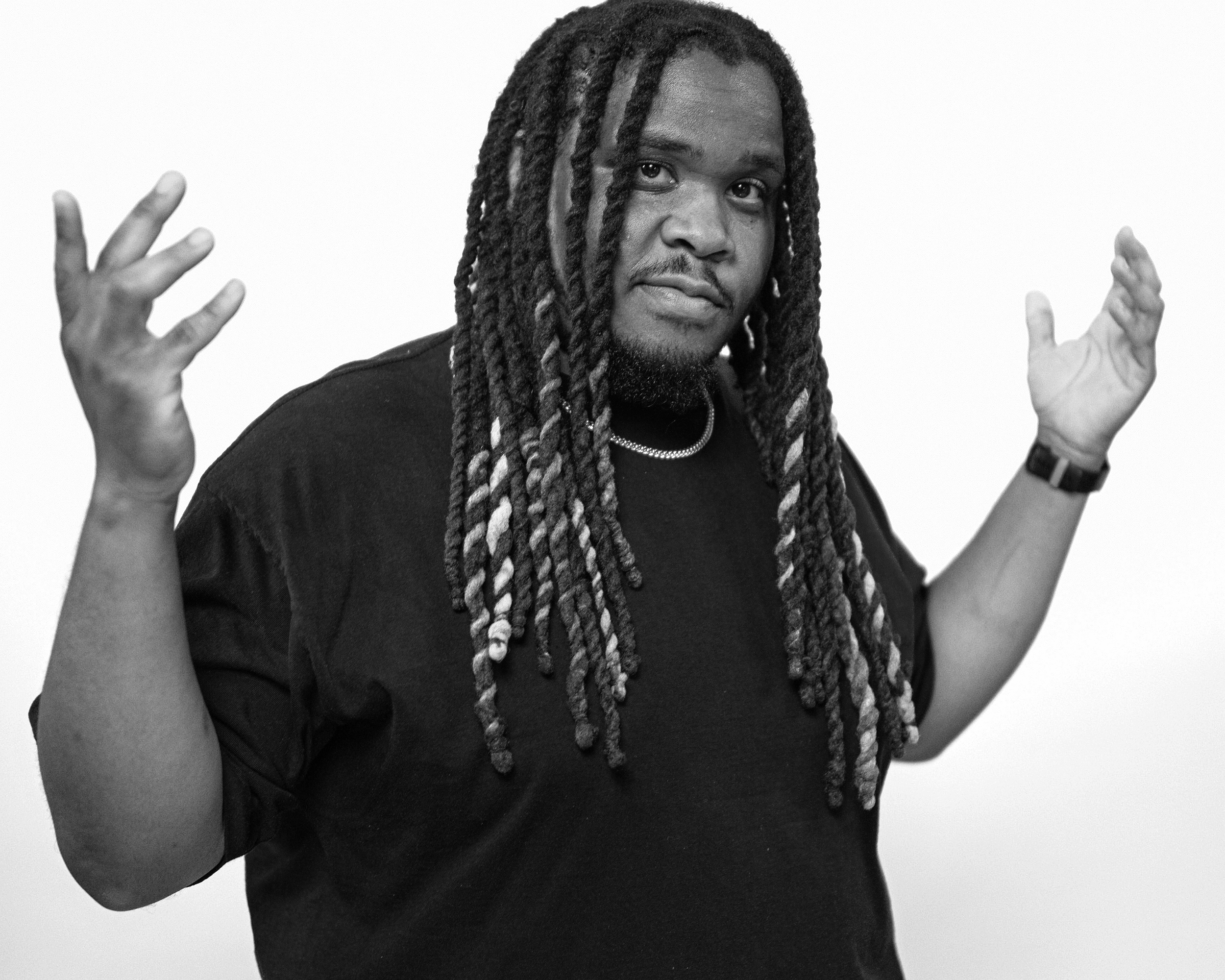 A man with long dreadlocks and a beard, wearing a black t-shirt, a silver chain, and a wristwatch, stands against a plain white background with his hands raised and palms facing outward.
