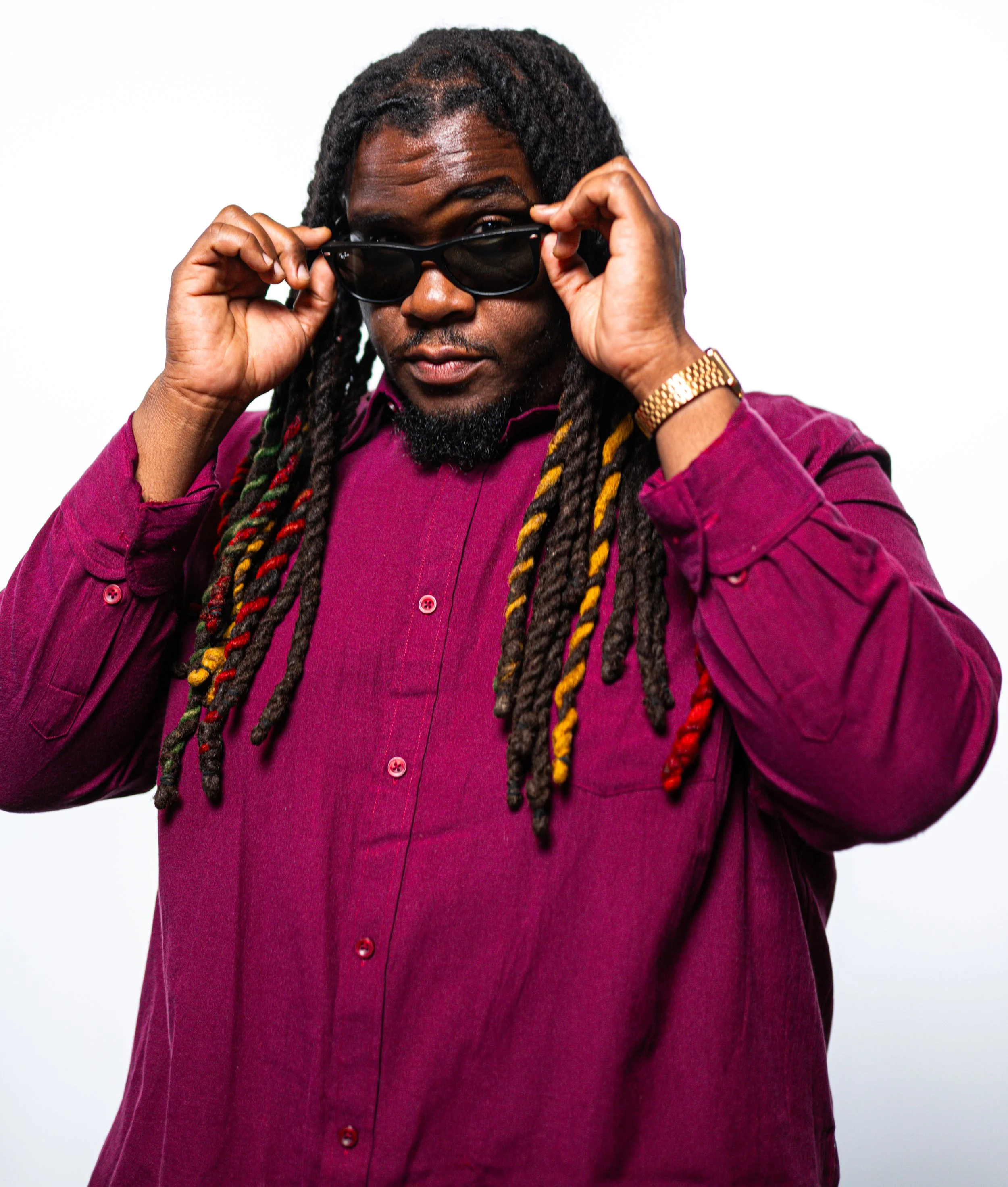 Mysta Crooks wearing sunglasses, a gold watch, and a maroon button-up shirt, holding his sunglasses with both hands, looking confident against a white background.