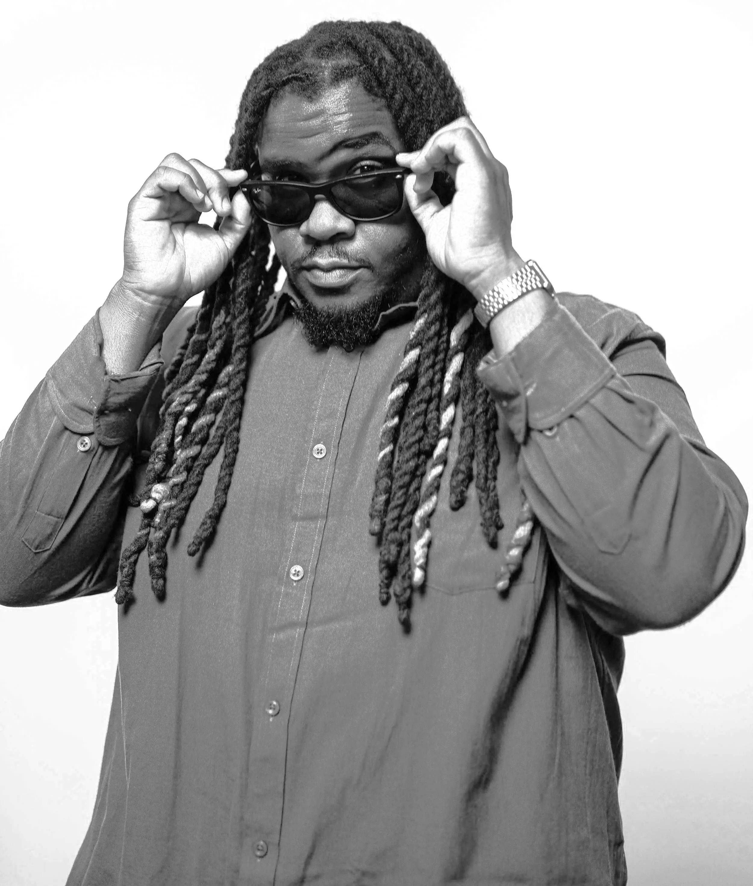 Mysta Crooks with long dreadlocks, beard, and sunglasses, holding sunglasses with both hands, looking at the camera, wearing a watch and a long-sleeve shirt, black and white photo.