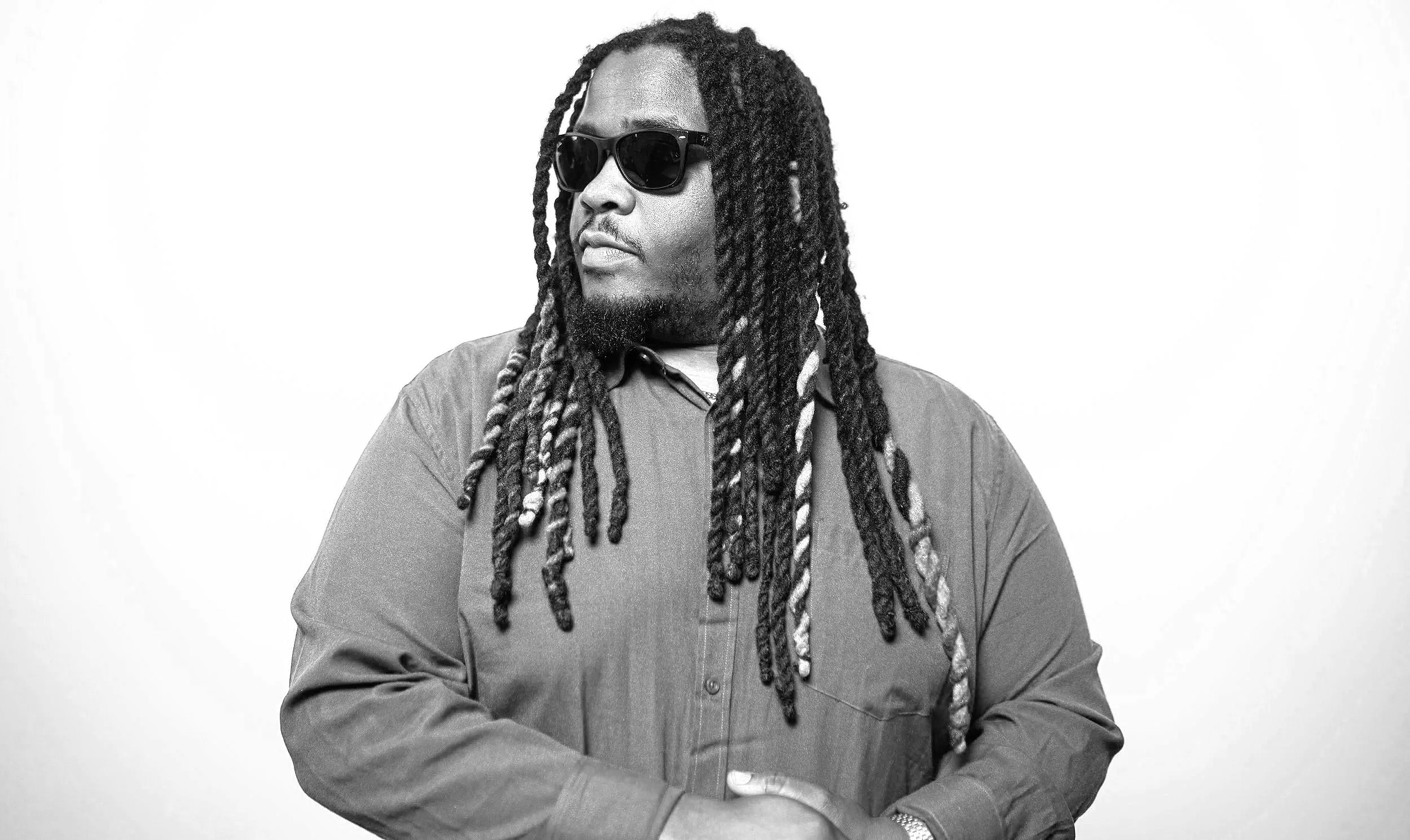 A Mysta Crooks wearing dark sunglasses and a button-up shirt, with long dreadlocks, standing against a plain white background, looking to his left.