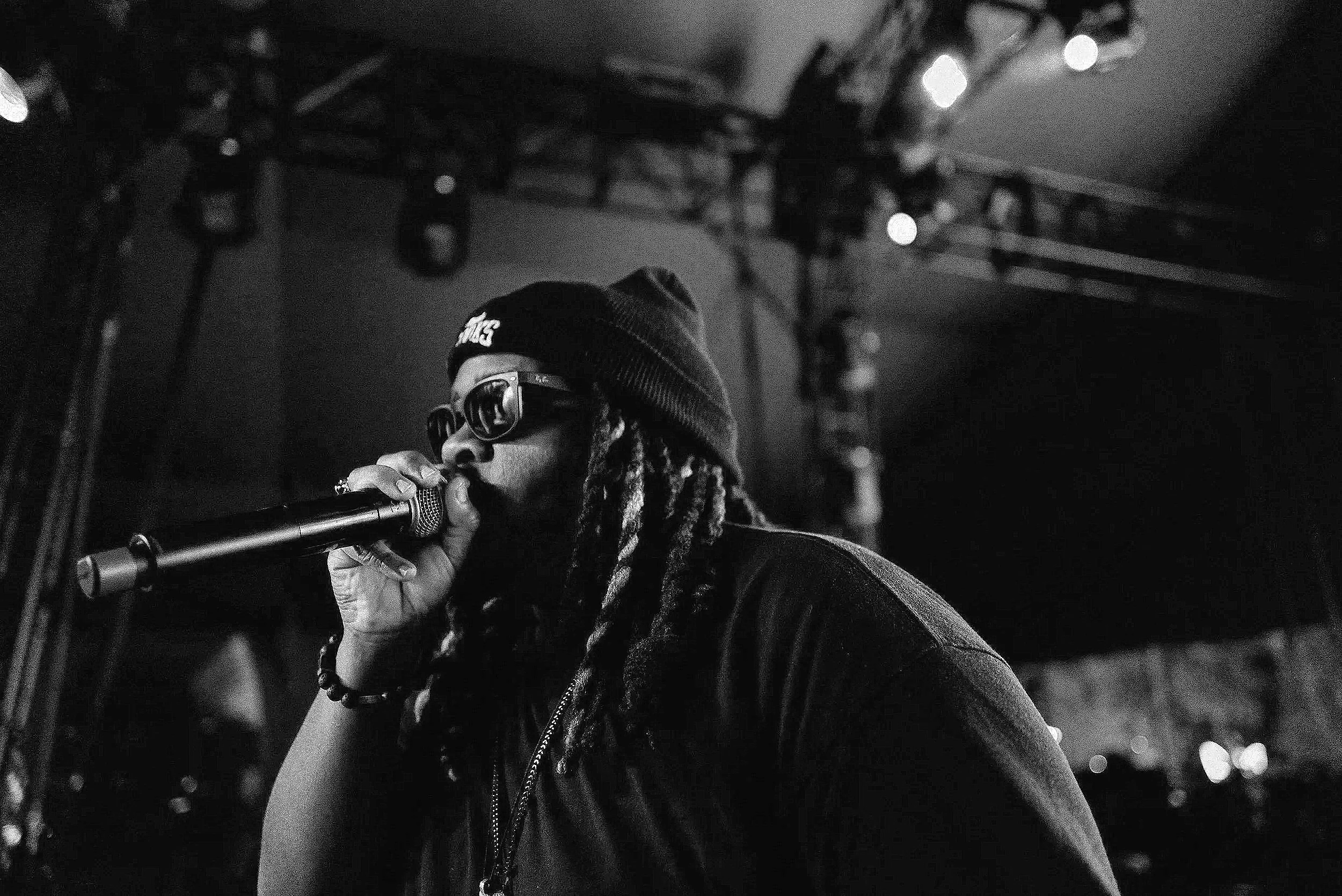 Mysta Crook with long dreadlocks wearing sunglasses and a beanie, MCing on stage at Coachella.