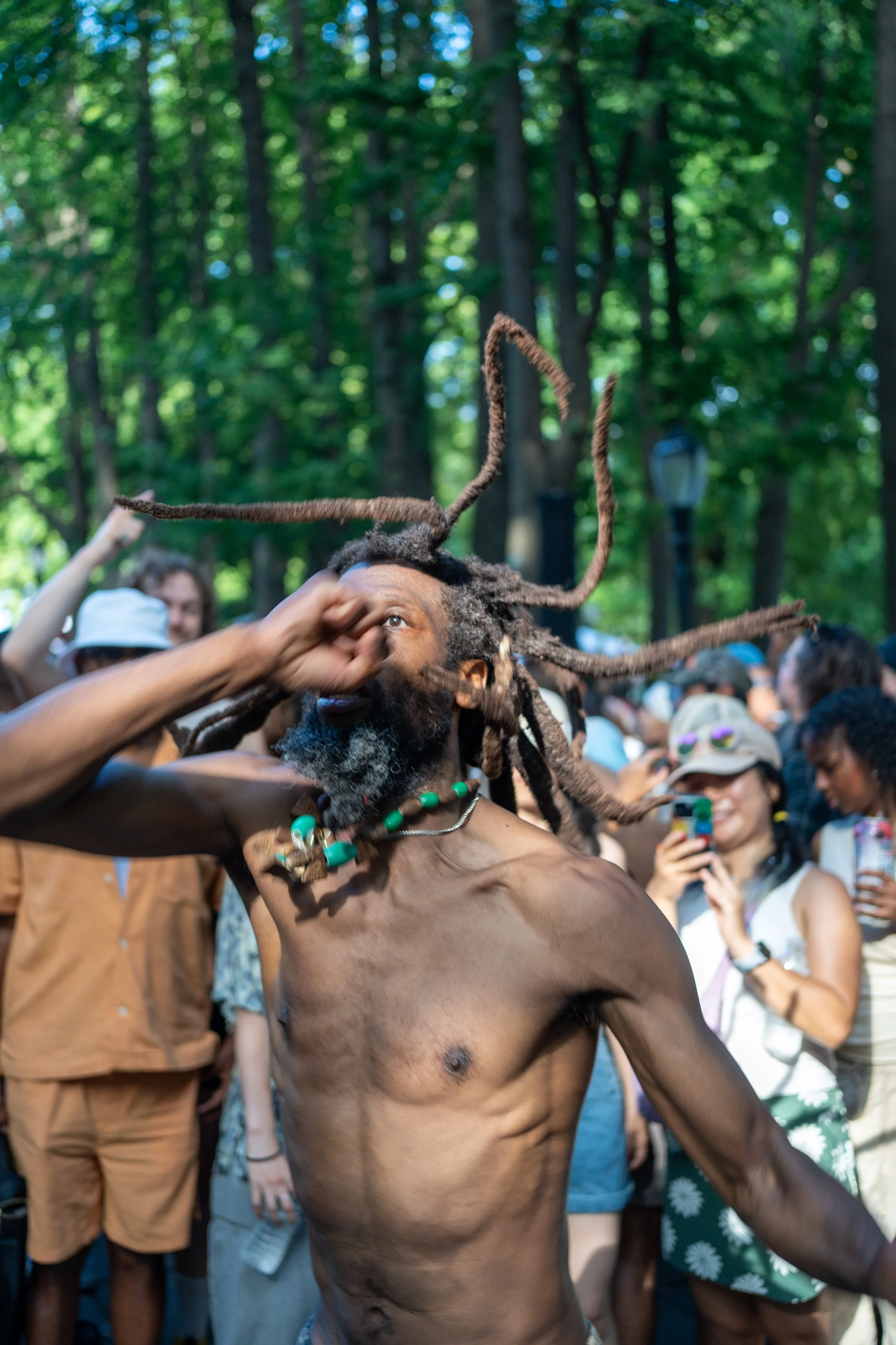 A shirtless man with dreadlocks and a beaded necklace dancing in front of a crowd outdoors, surrounded by trees.