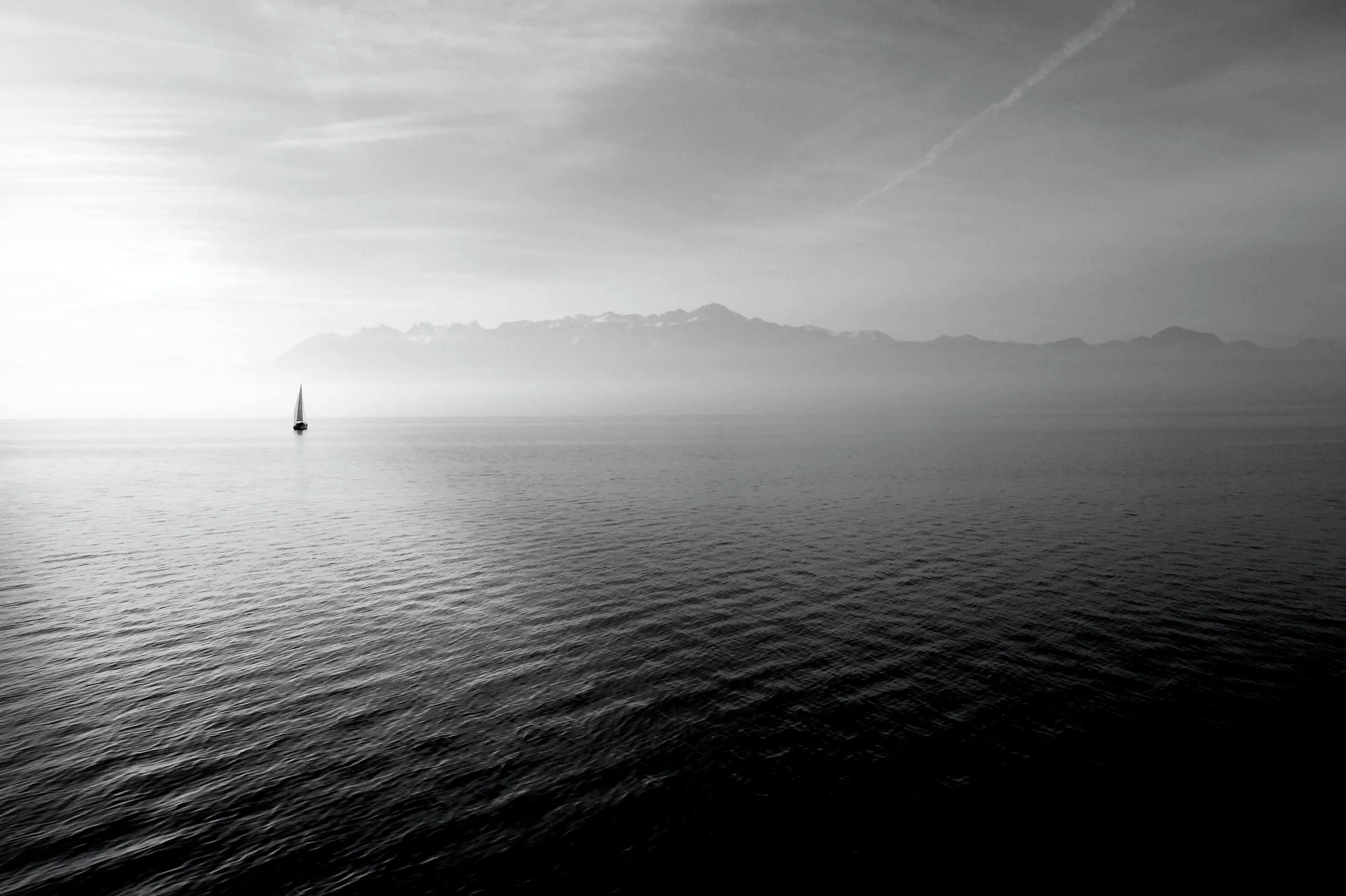 Black and white photo of the still ocean with clouds behind a lone sailboat.