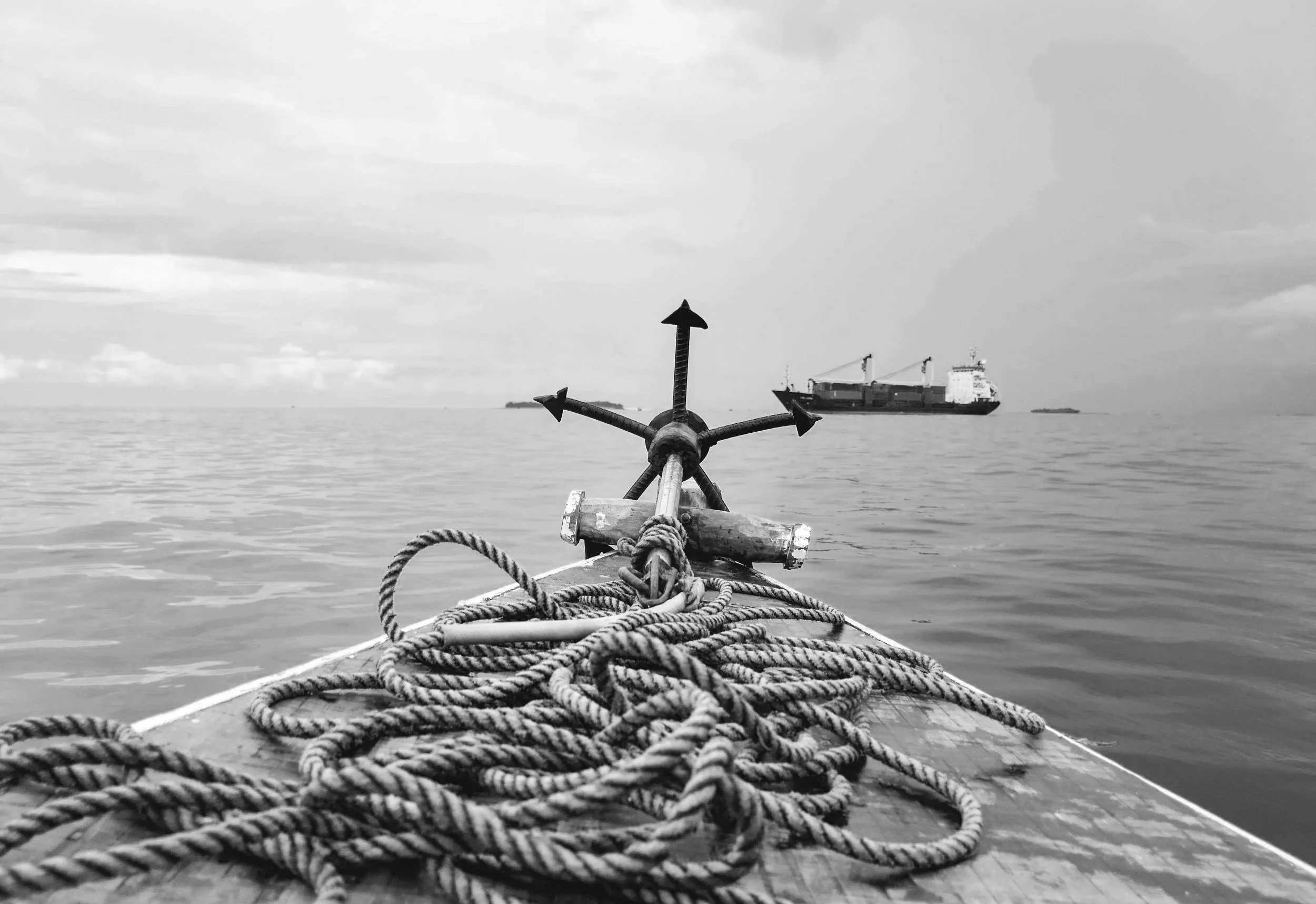 Black and white photo of a 5 point anchor and its rope sitting on a dock pointing at a ocean freighter in the distance