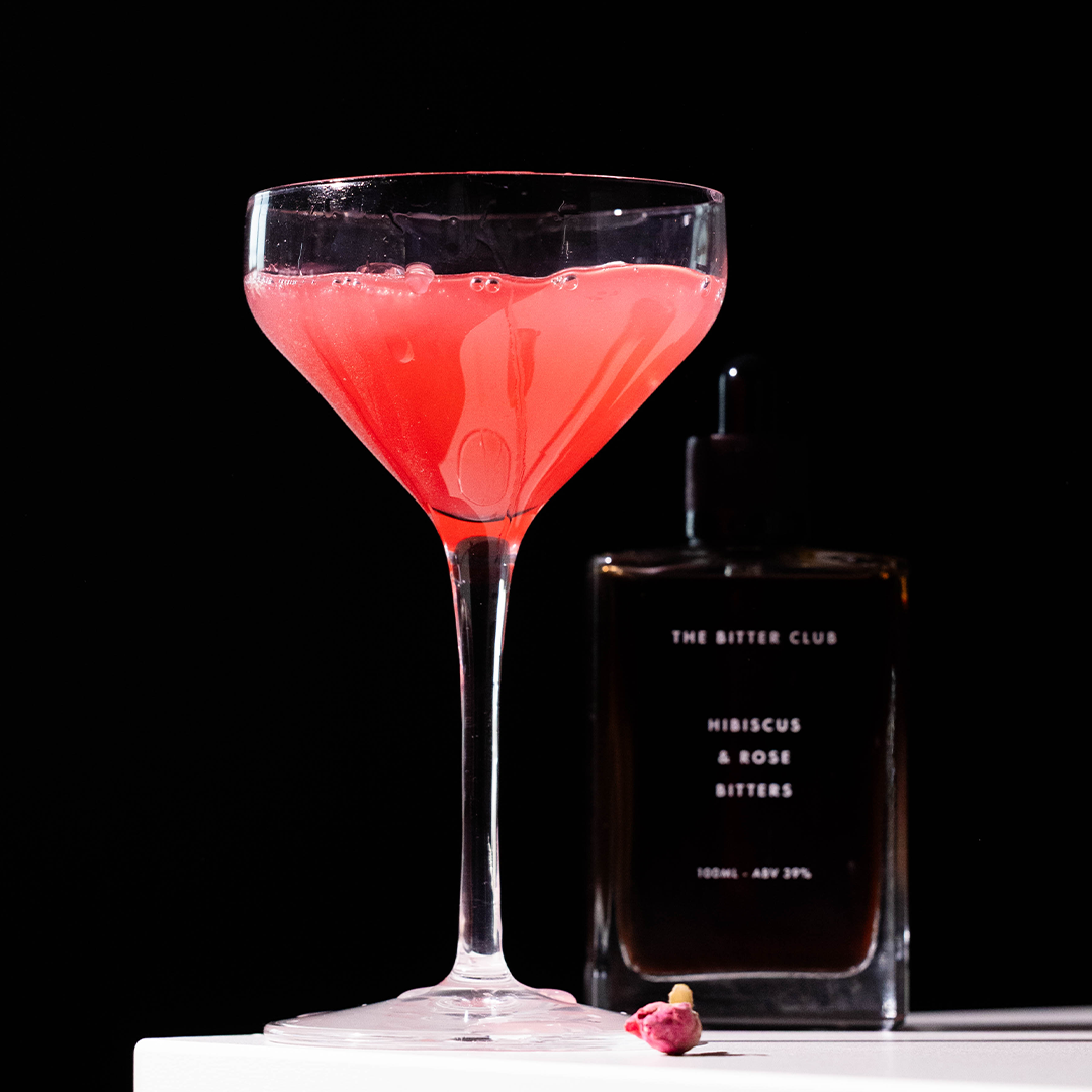 A pink cocktail in a martini glass with bubbles, next to a bottle labeled 'The Bitter Club Hibiscus & Rose Bitters' against a black background.
