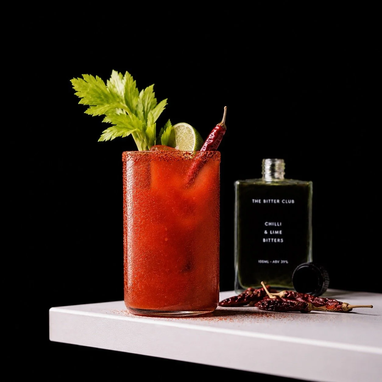 A cocktail in a tall glass with condensation, garnished with celery, lime, and dried red chili peppers, placed on a white surface with a black background. A bottle of bitters labeled 'Chilli & Lime Bitters' is in the background.