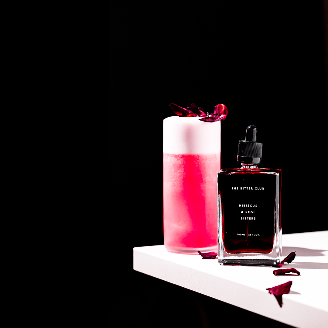 A bottle of hibiscus and rose bitters labeled 'The Bitter Club' next to a pink and white layered cocktail with flower garnish, on a white surface against a black background.