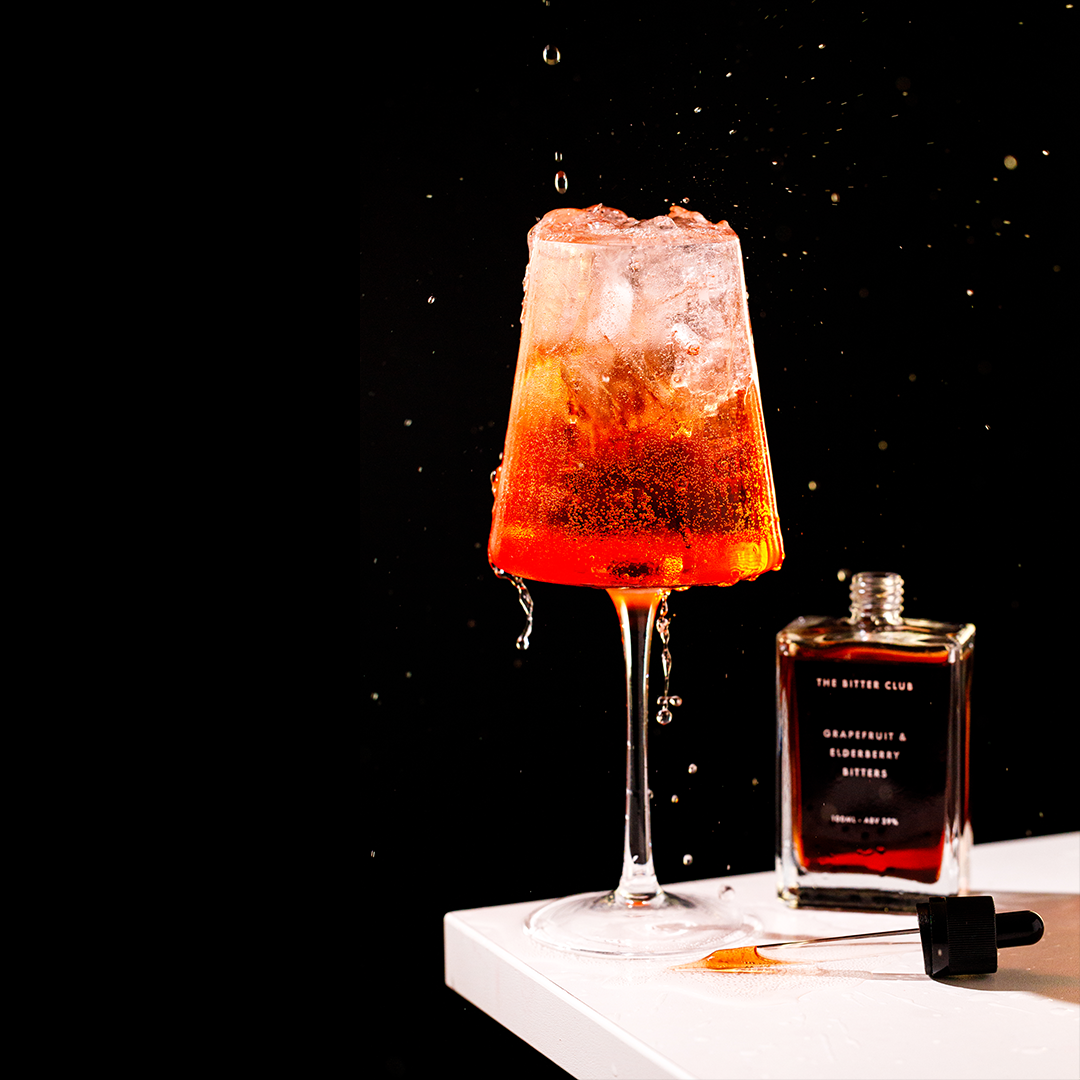 A cocktail glass filled with a red-orange beverage and ice cubes, with droplets falling from the drink against a black background. A bottle labeled 'Grapefruit & Elderberry Bitters' and a dropper lie on a white surface next to the glass.