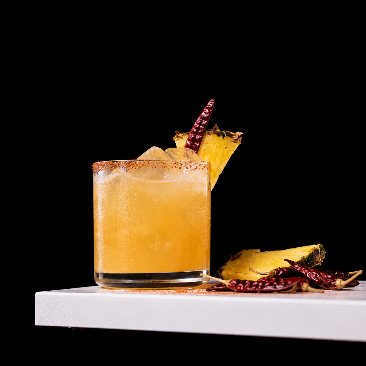A glass of yellow cocktail with ice, garnished with pineapple and dried red chili peppers, on a white surface against a black background.