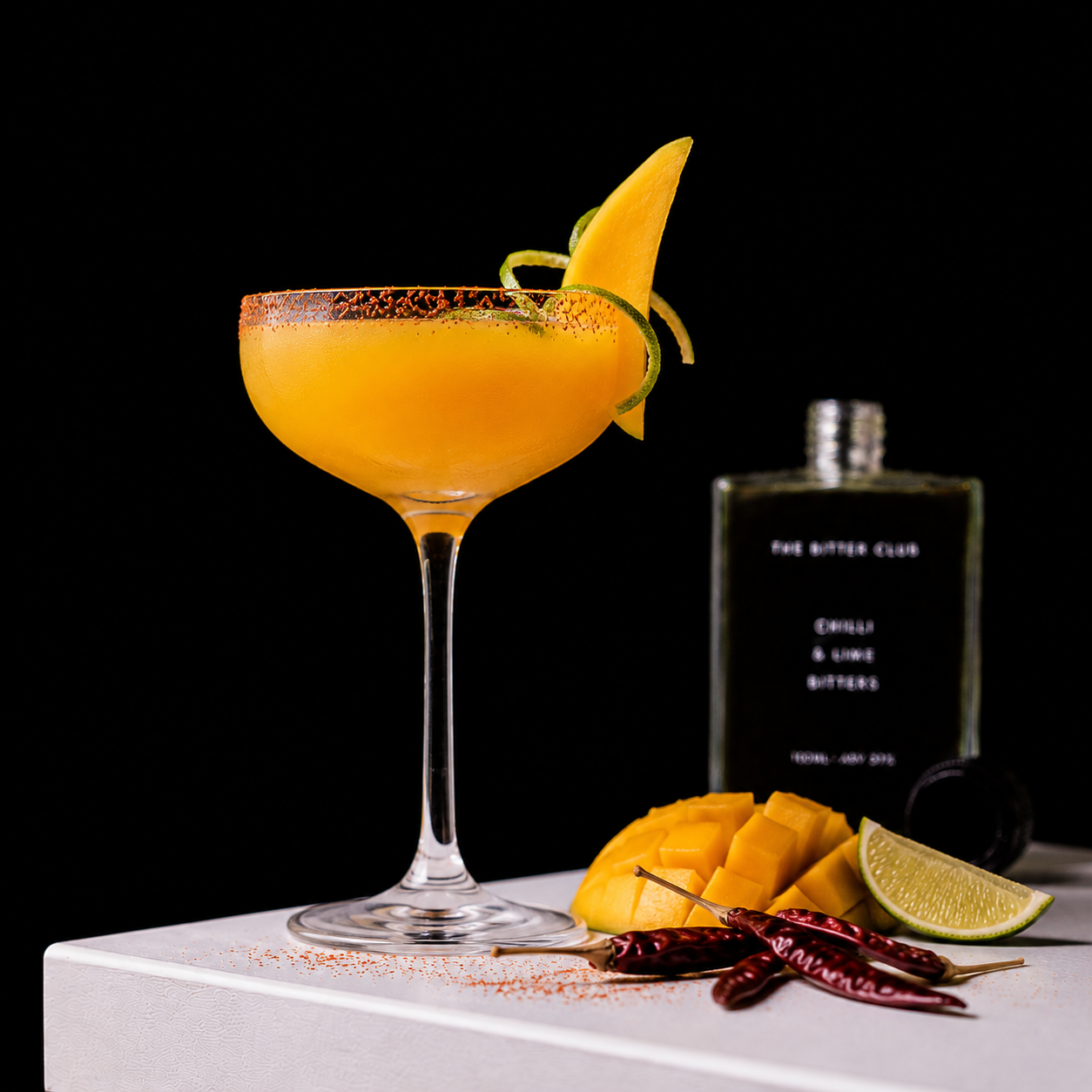 A cocktail in a coupe glass garnished with a lemon wedge and lime twist, served with cubes of mango, dried red chili peppers, and a lime wedge on a white surface, with a bottle labeled 'The Bitter Club' in the background.