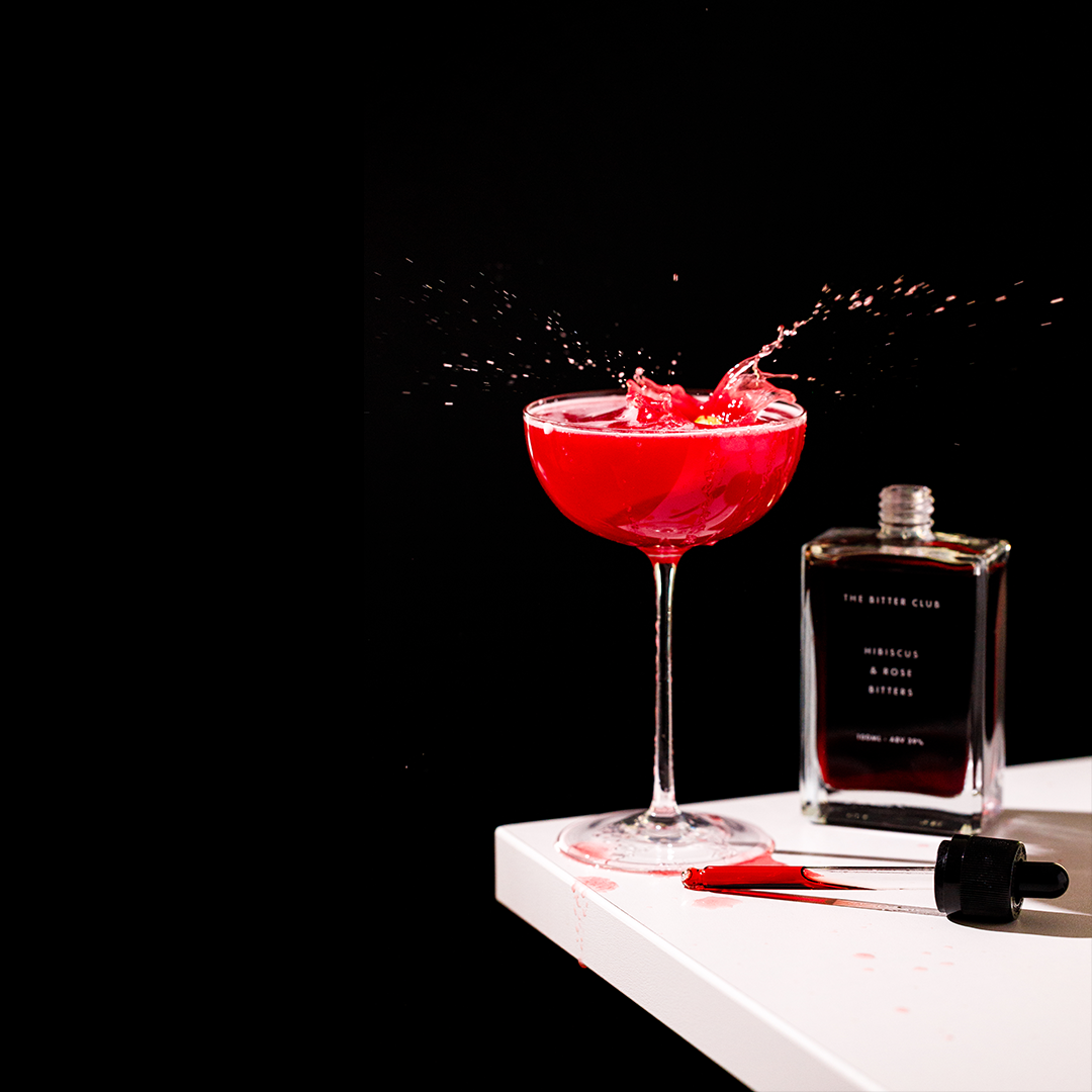 A red cocktail in a coupe glass with splash effects, a glass bottle labeled 'The Bitter Club,' and a dropper on a white surface against a black background.