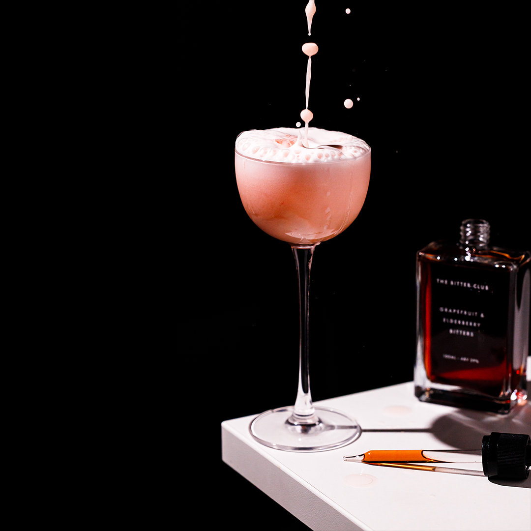 A pink cocktail in a coupe glass with a foamy top, with liquid pouring into it, placed on a white surface next to a bottle of dark liquor and a pen.