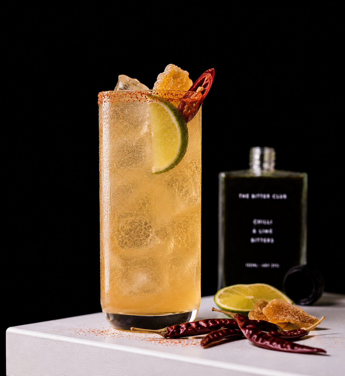 A tall glass of a yellow cocktail garnished with a slice of lime and red chili pepper, with ice cubes and condensation on the outside, placed on a white surface. There are dried red chili peppers, a lemon wedge, and a piece of candied ginger nearby, with a dark background and a blurred bottle in the background.