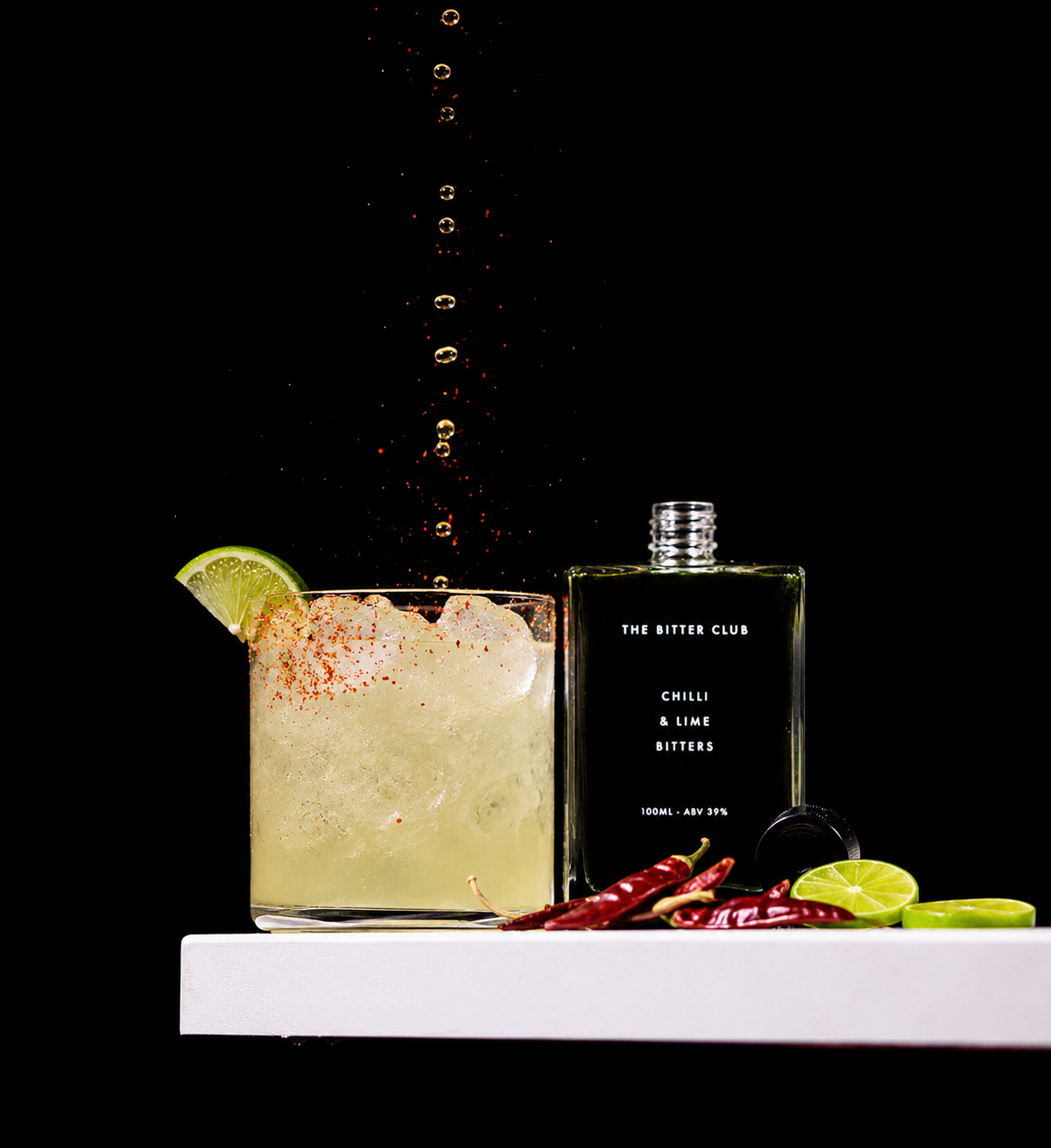 A glass of cocktail with lime wedge on the rim, topped with chili salt, ice, and red chili peppers, next to a bottle of bitters labeled 'The Bitter Club Chilli & Lime Bitters', on a white surface against a black background.