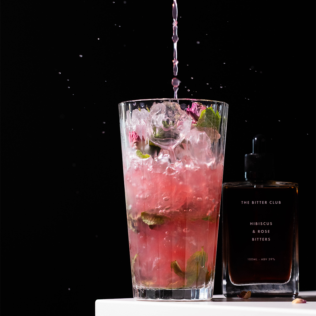 Pink cocktail with ice, mint leaves, and ice cubes in a tall glass, with a bottle of hibiscus and rose bitters nearby, on a white surface against a black background.