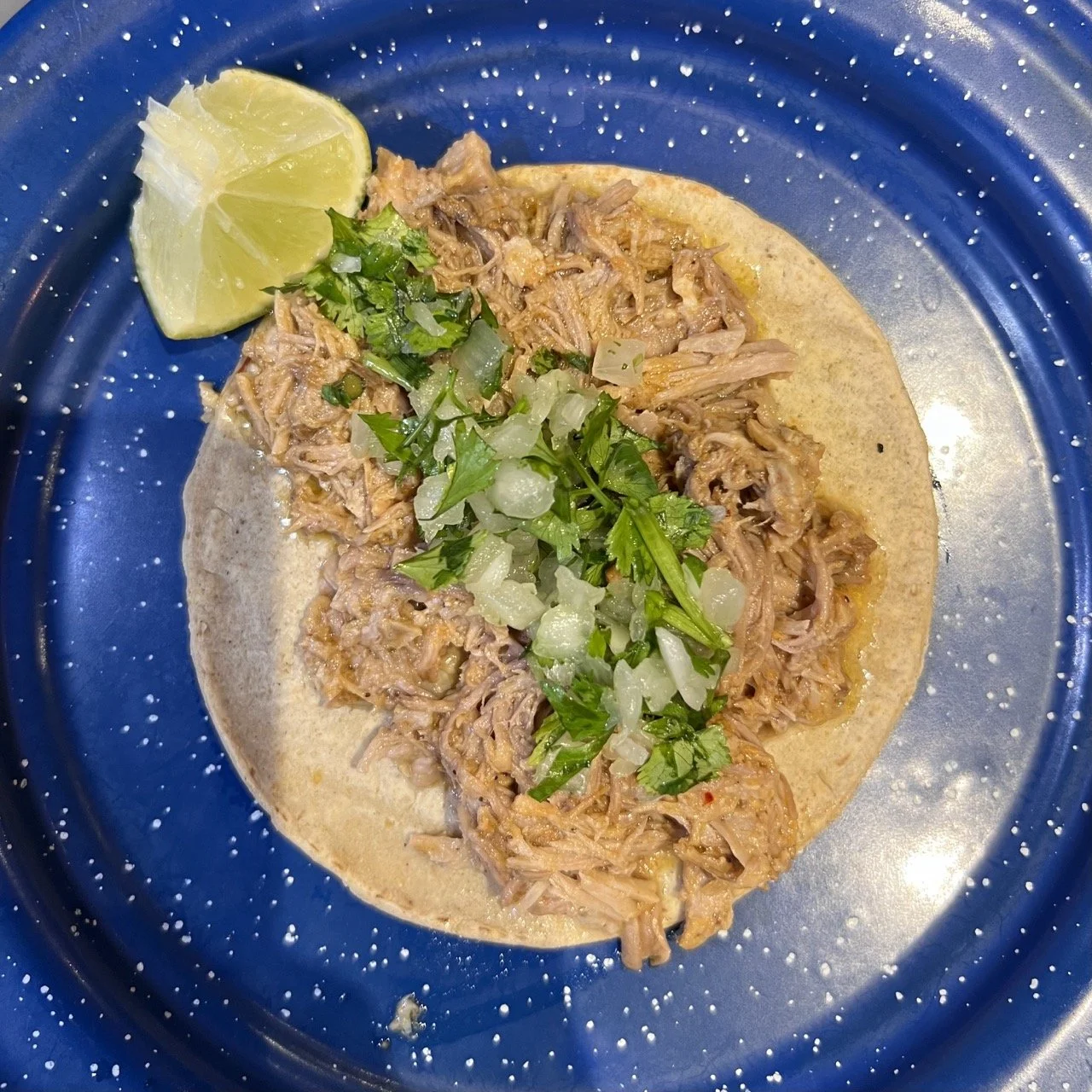 Taco avec viande effilochée, oignons, coriandre et quartier de citron vert sur une assiette bleue.