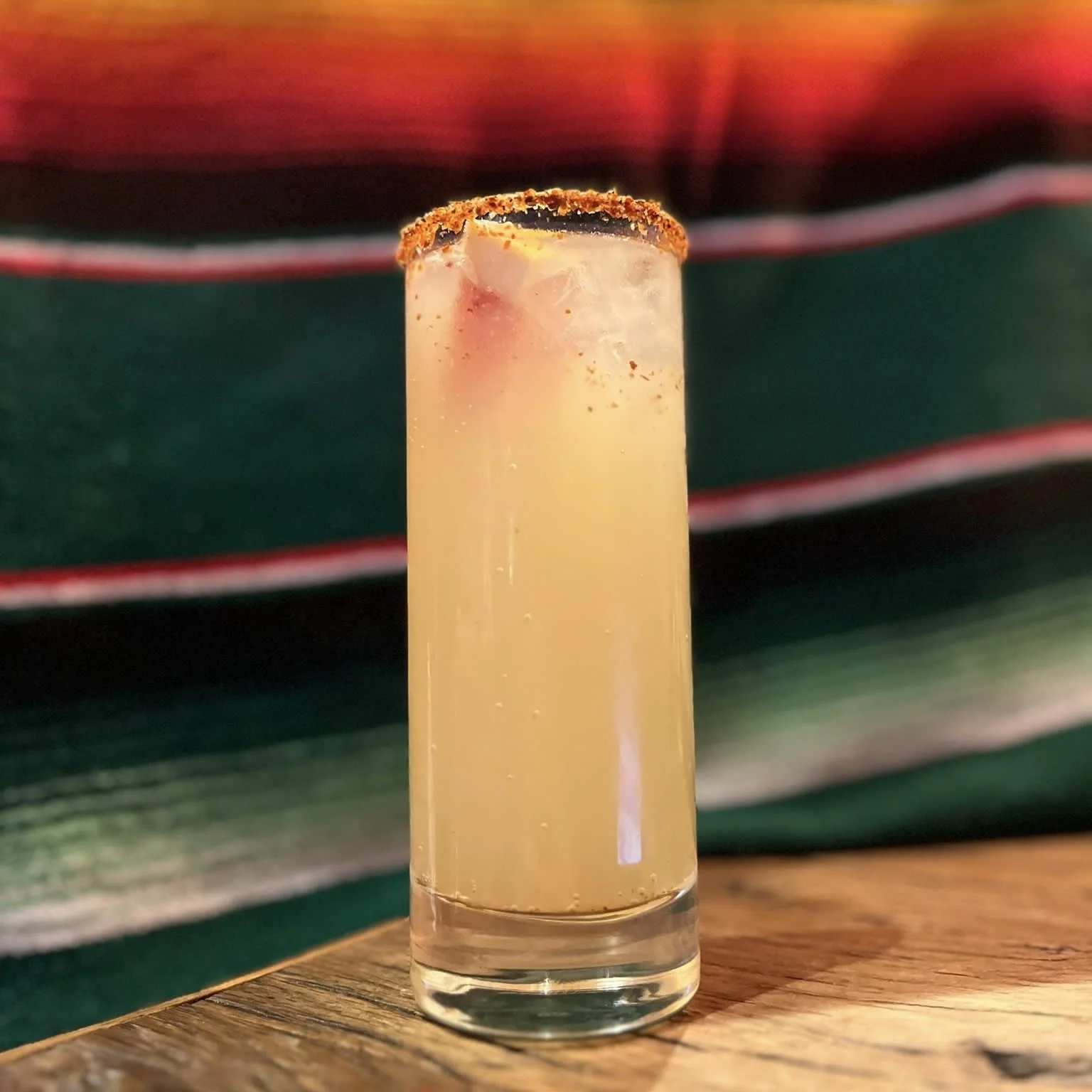 Un cocktail clair avec des glaçons dans un verre à long pied, garni d'un bord épicé. En arrière-plan, un tissu rayé coloré.