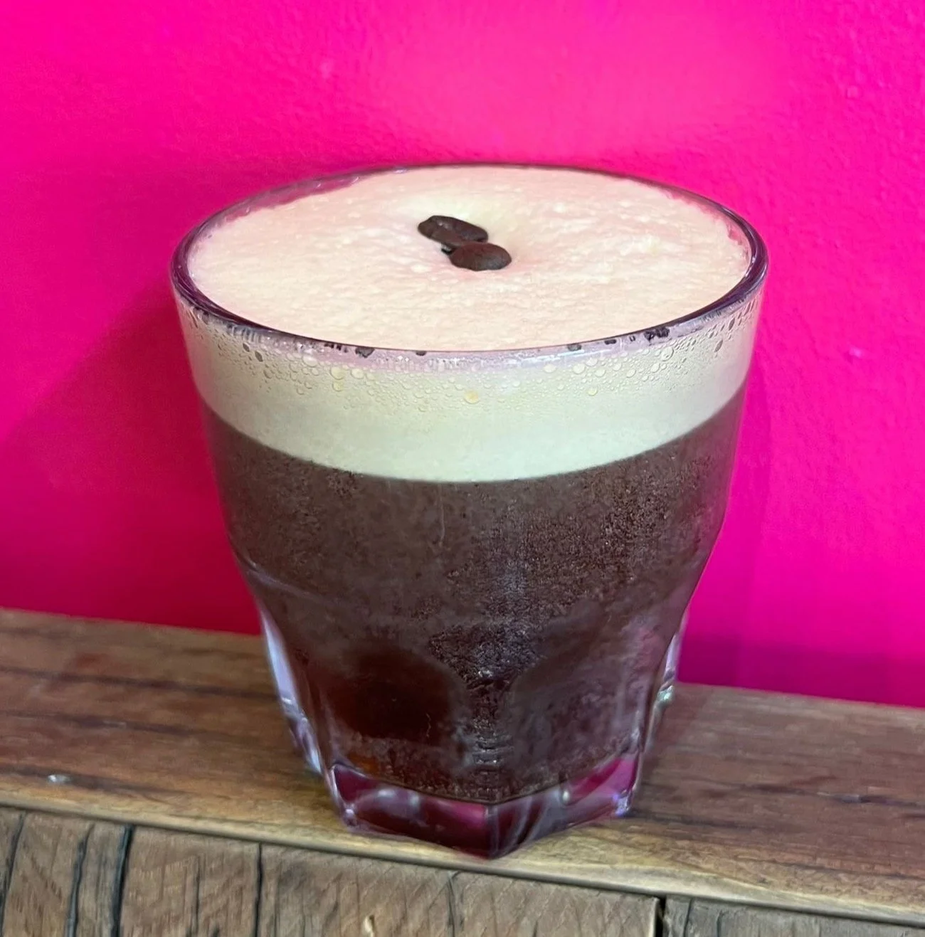 Verre de cocktail sur une table en bois, avec mousse crémeuse et grains de café en décoration, fond rose.