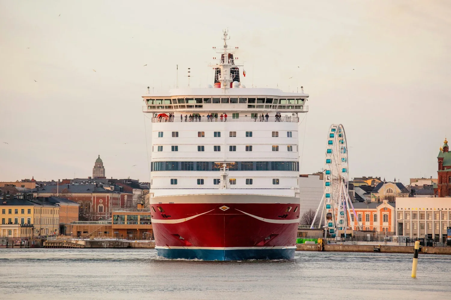 Large cruise ship sailing in a harbor with a city skyline, including a Ferris wheel, in the background.