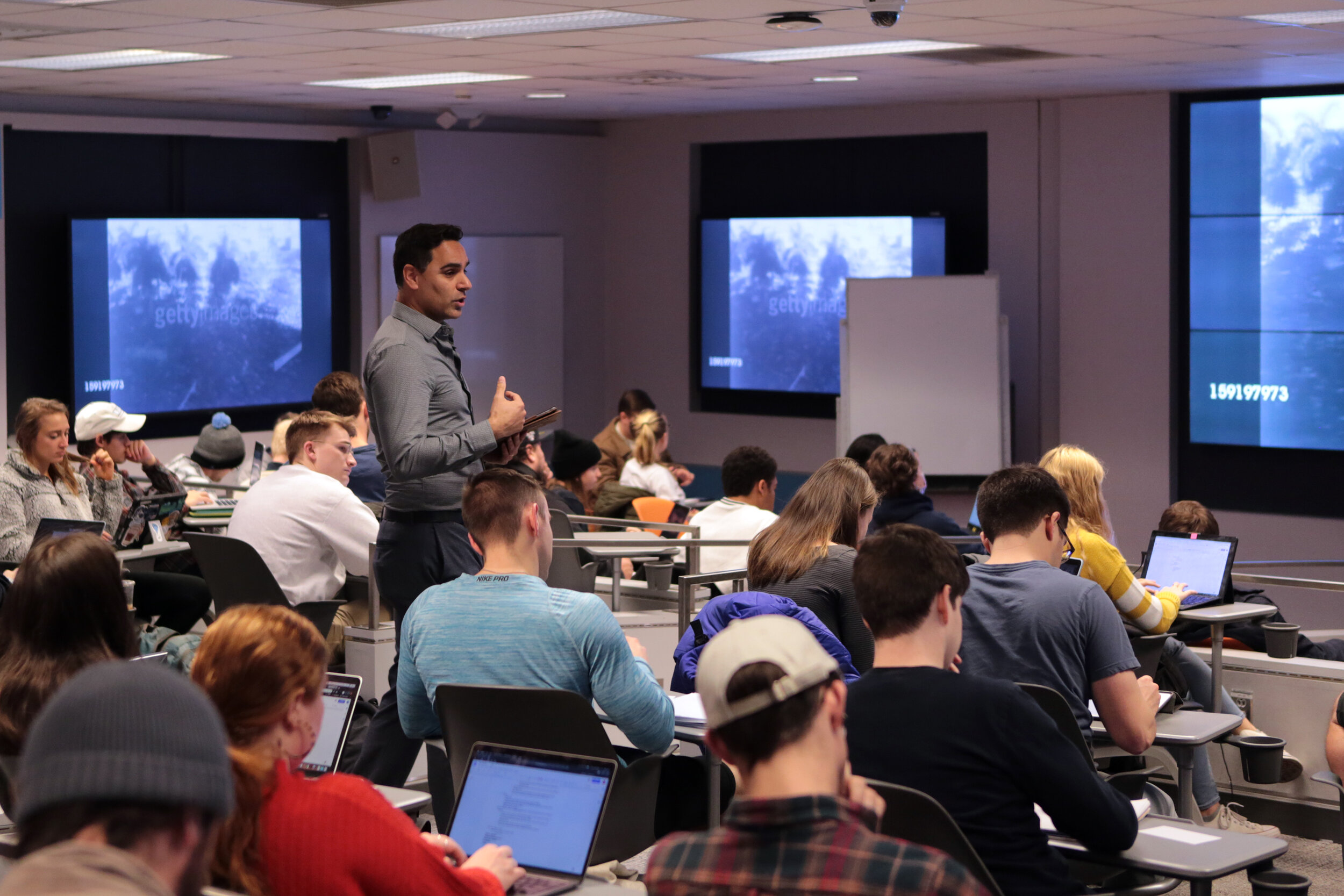Professor La Serna stands in the middle of a classroom of students gesturing and seemingly mid-sentence. Students in seats around him take notes and we can see three screens projecting documentary footage around the classroom.