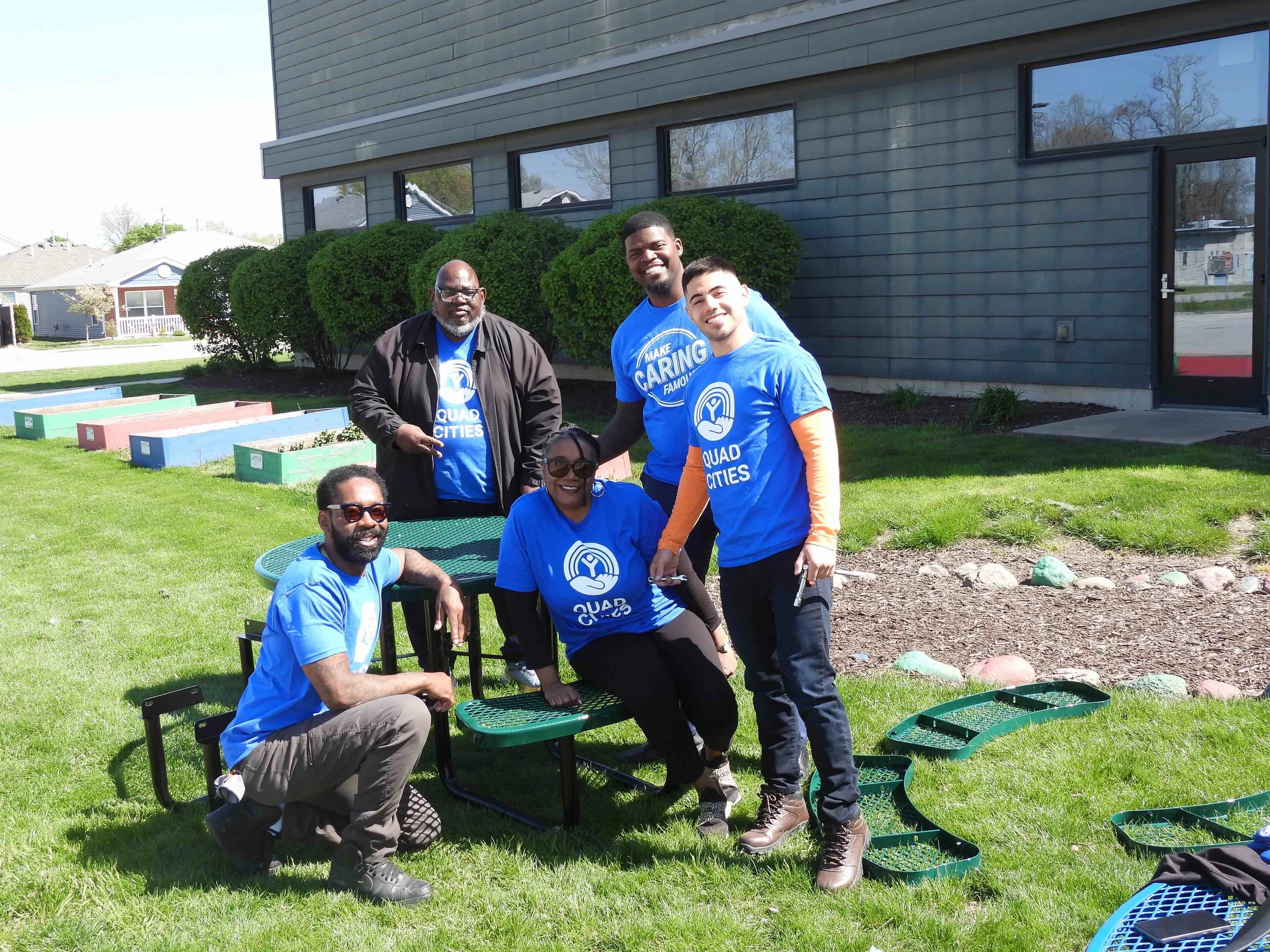 A group of five people wearing blue "Quad Cities" t-shirts are gathered outdoors on a sunny day, smiling and posing near a building with large windows. Some are sitting and others are standing on a grassy area, near some colorful raised garden beds and patio furniture.