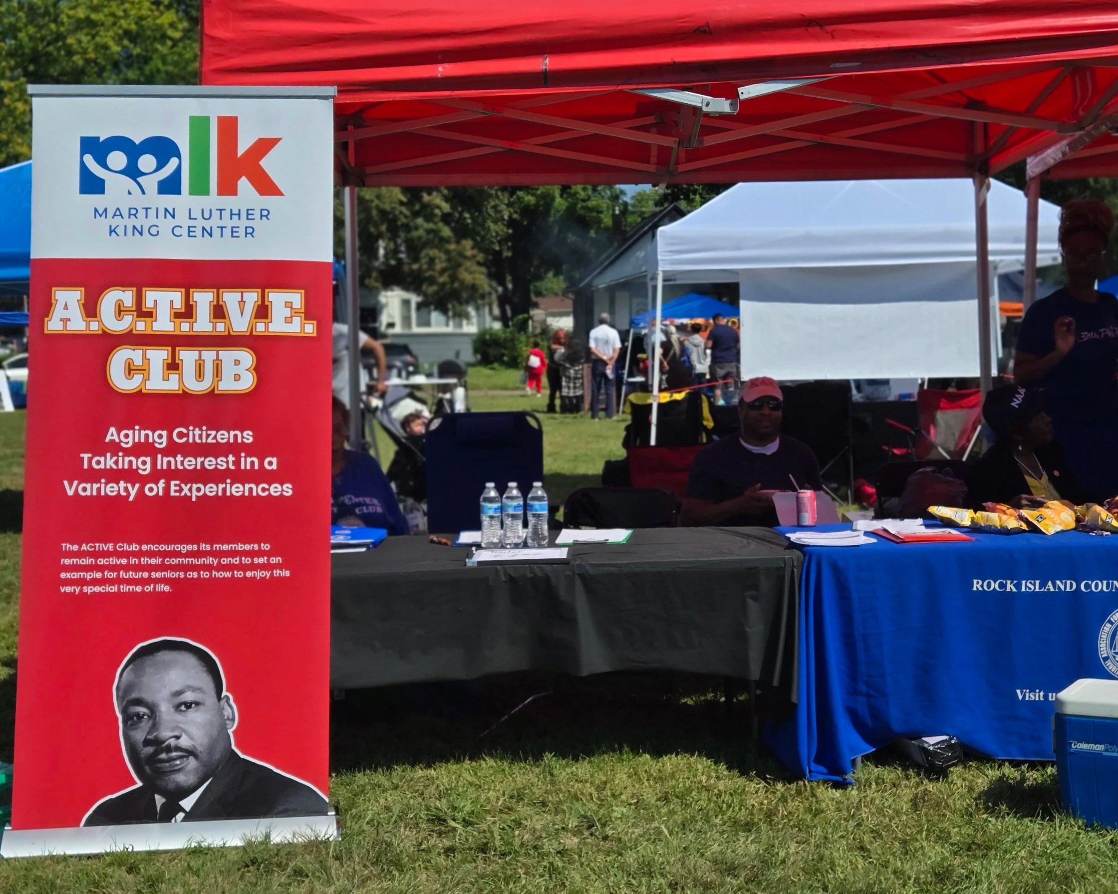Outdoor community event with tents and banners, featuring a red banner with a black and white portrait of Martin Luther King Jr. and a logo of a blue face with a white escutcheon. The background shows several people, some wearing red and other brightly colored clothing, attending the event on a grassy area under a blue sky.