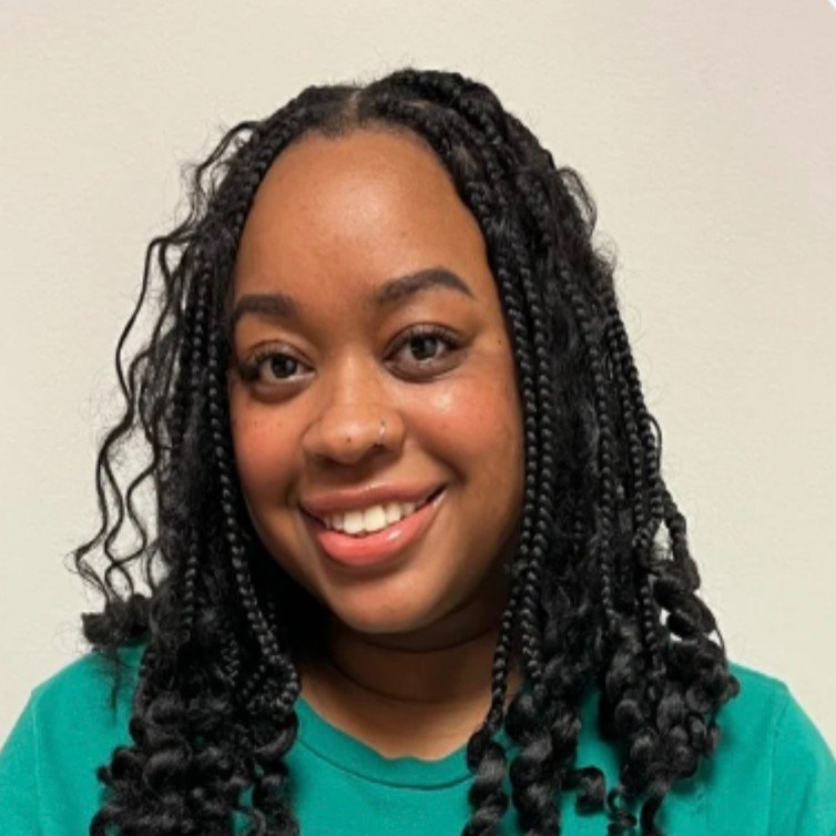 A young woman with dark, curly, twisted hair wearing a teal shirt and smiling at the camera against a plain background.