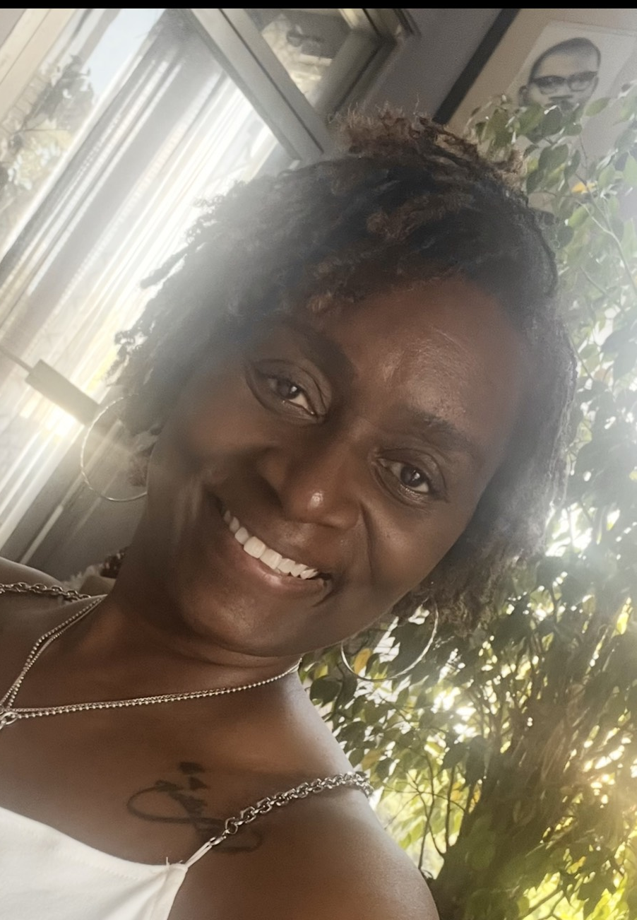 The image depicts a person smiling at the camera. They have short, curly hair and are wearing hoop earrings and a white top with spaghetti straps and a chain detail.