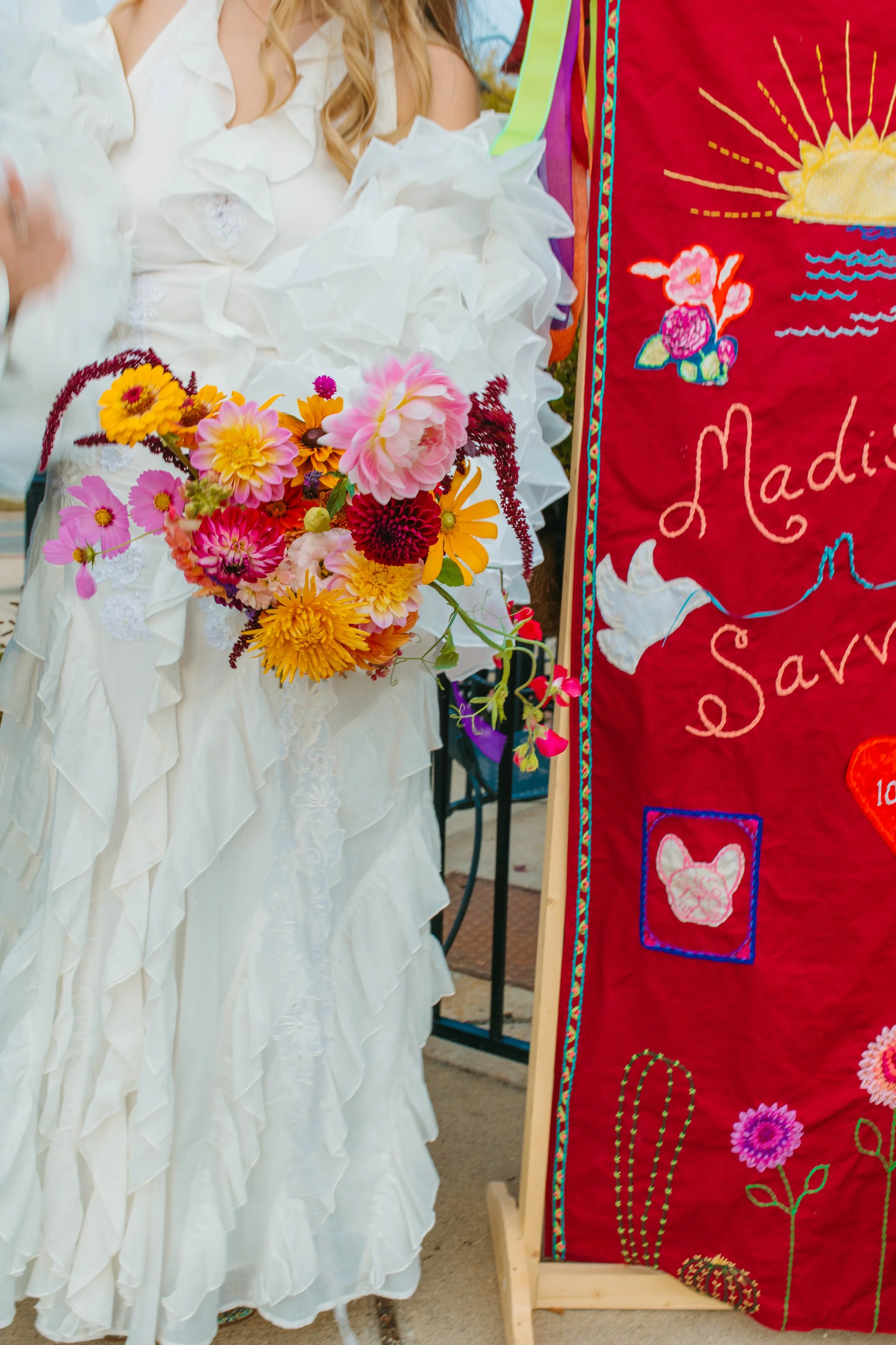 A person wearing a white, ruffled dress holds a bouquet of colorful flowers including pink, yellow, and red blooms. Next to them is a red fabric with embroidered flowers, a sun, and the word 'Madison' visible.