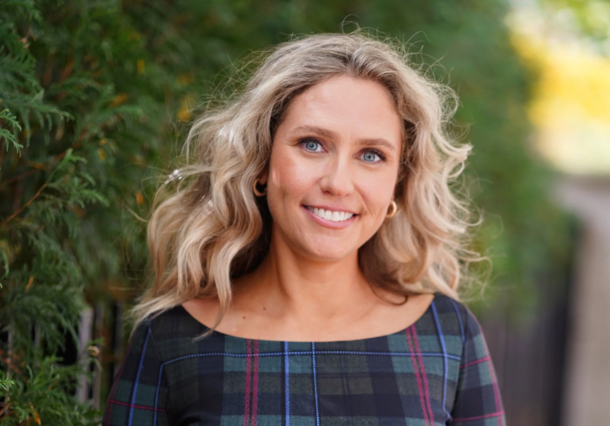 A woman with wavy blonde hair, blue eyes, and hoop earrings outdoors, smiling, wearing a plaid top, with greenery in the background.
