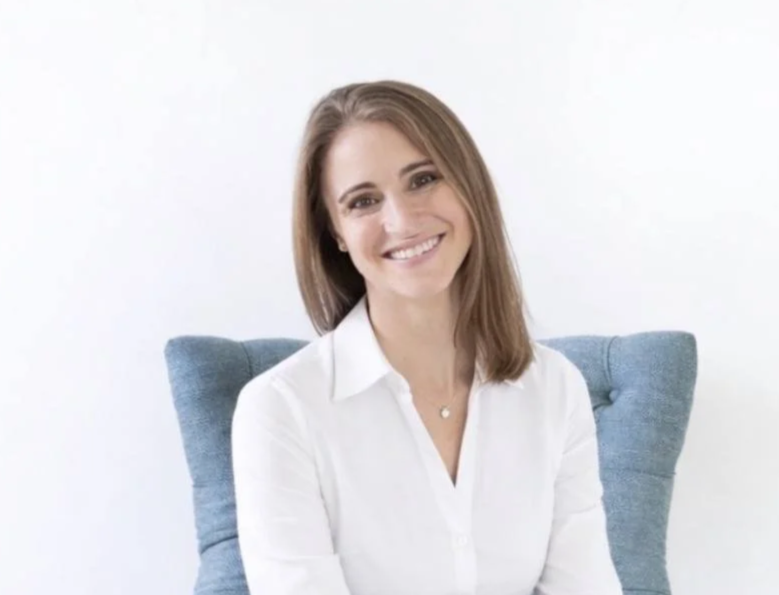 A woman with shoulder-length brown hair, wearing a white blouse, smiling while sitting in a light blue armchair.