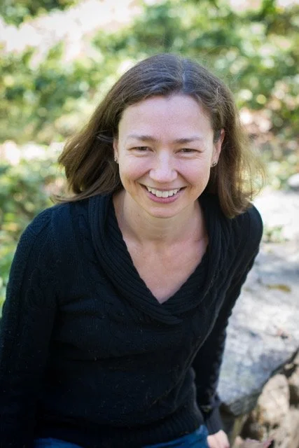 Smiling woman with shoulder-length brown hair, wearing a black top, outdoors with green foliage in the background.