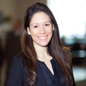 Portrait of a woman with long brown hair smiling, wearing a dark blazer and navy top, indoors with blurred background.