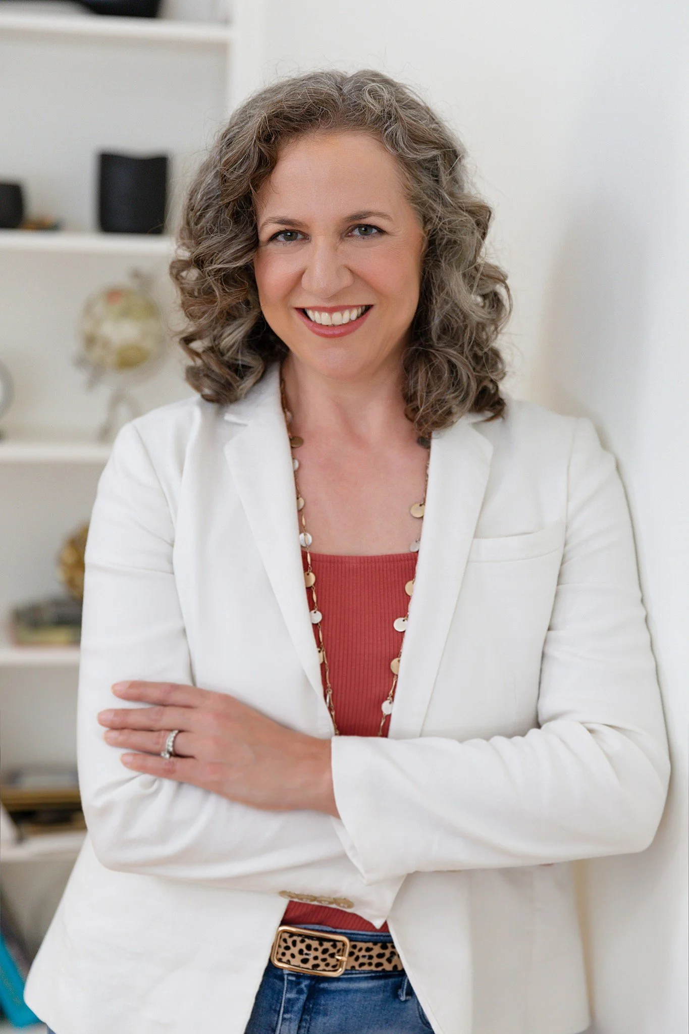 A woman with curly gray hair smiling, wearing a white blazer, red top, leopard print belt, and jewelry, standing in a home or office setting.