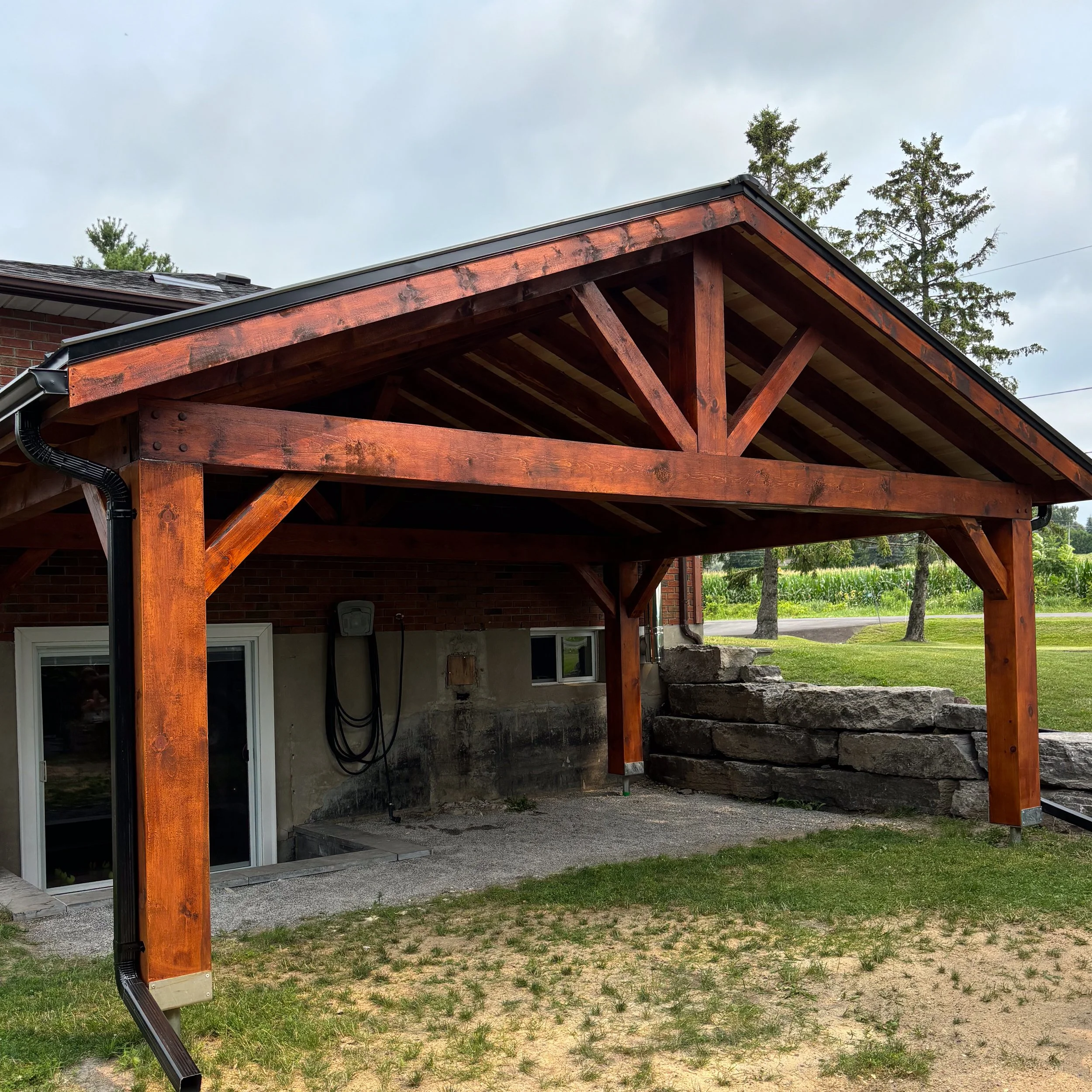 A wooden deck with a roof structure attached to the back of a brick house, with rocks and grass in the yard and trees in the background.