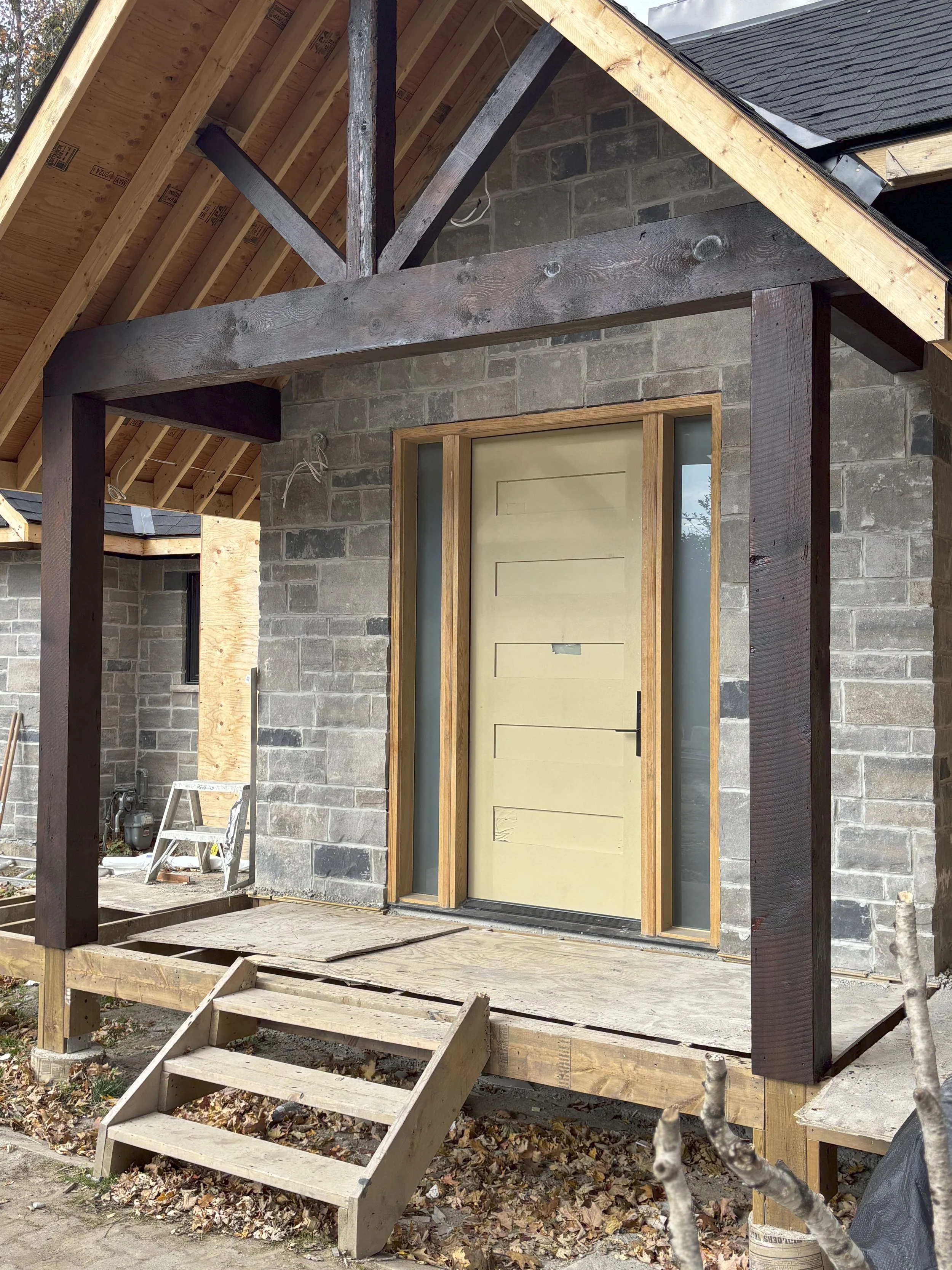 Front porch under construction of a house with a yellow front door and grey brick walls, wooden stairs, and a wooden frame around the door.
