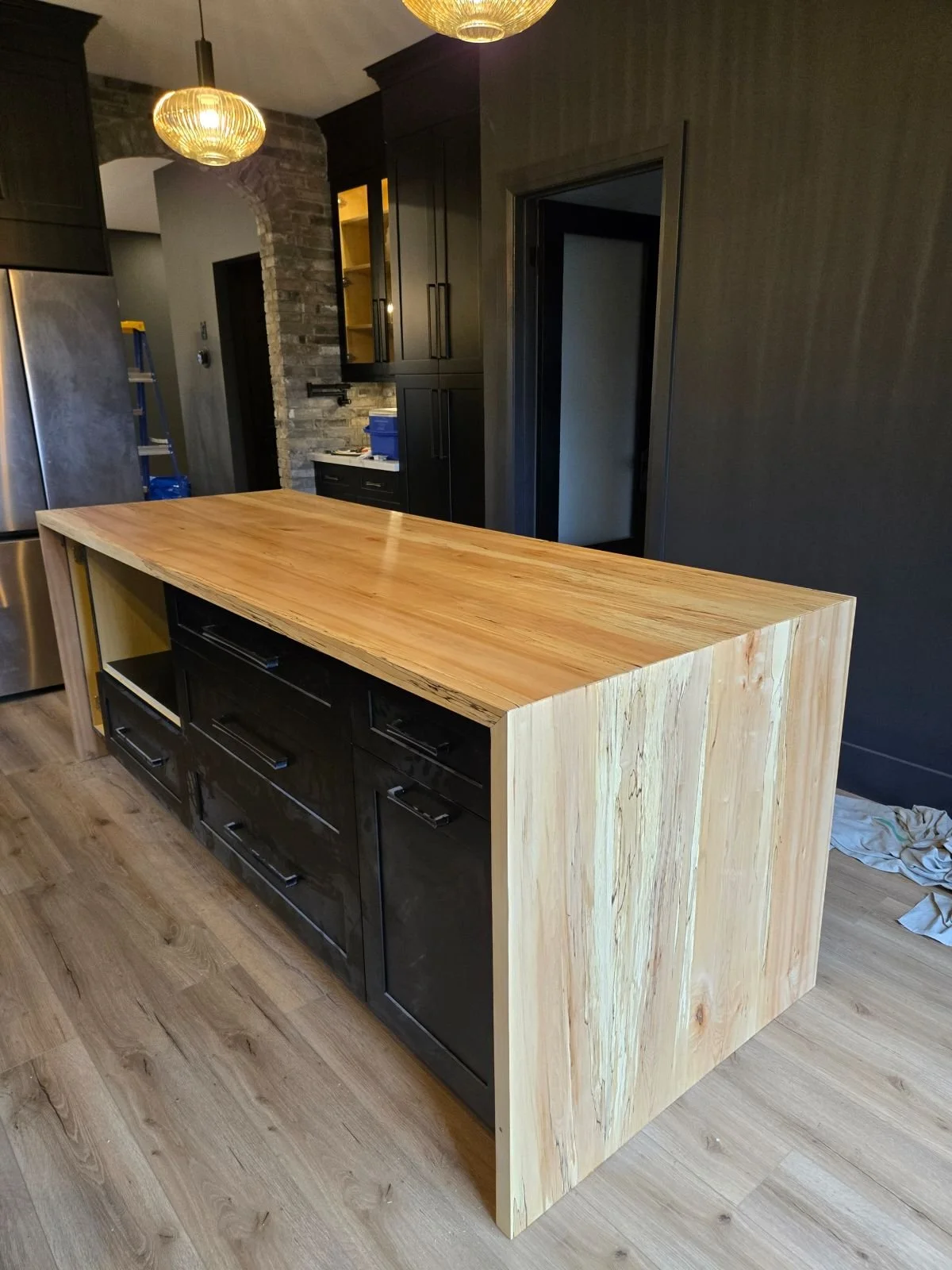 A modern kitchen island with a natural wood top and black drawers and cabinets, set in a kitchen with dark cabinets, stainless steel appliances, and a brick archway. Custom wooden counter top, durham region - kawartha lakes - northumberland county