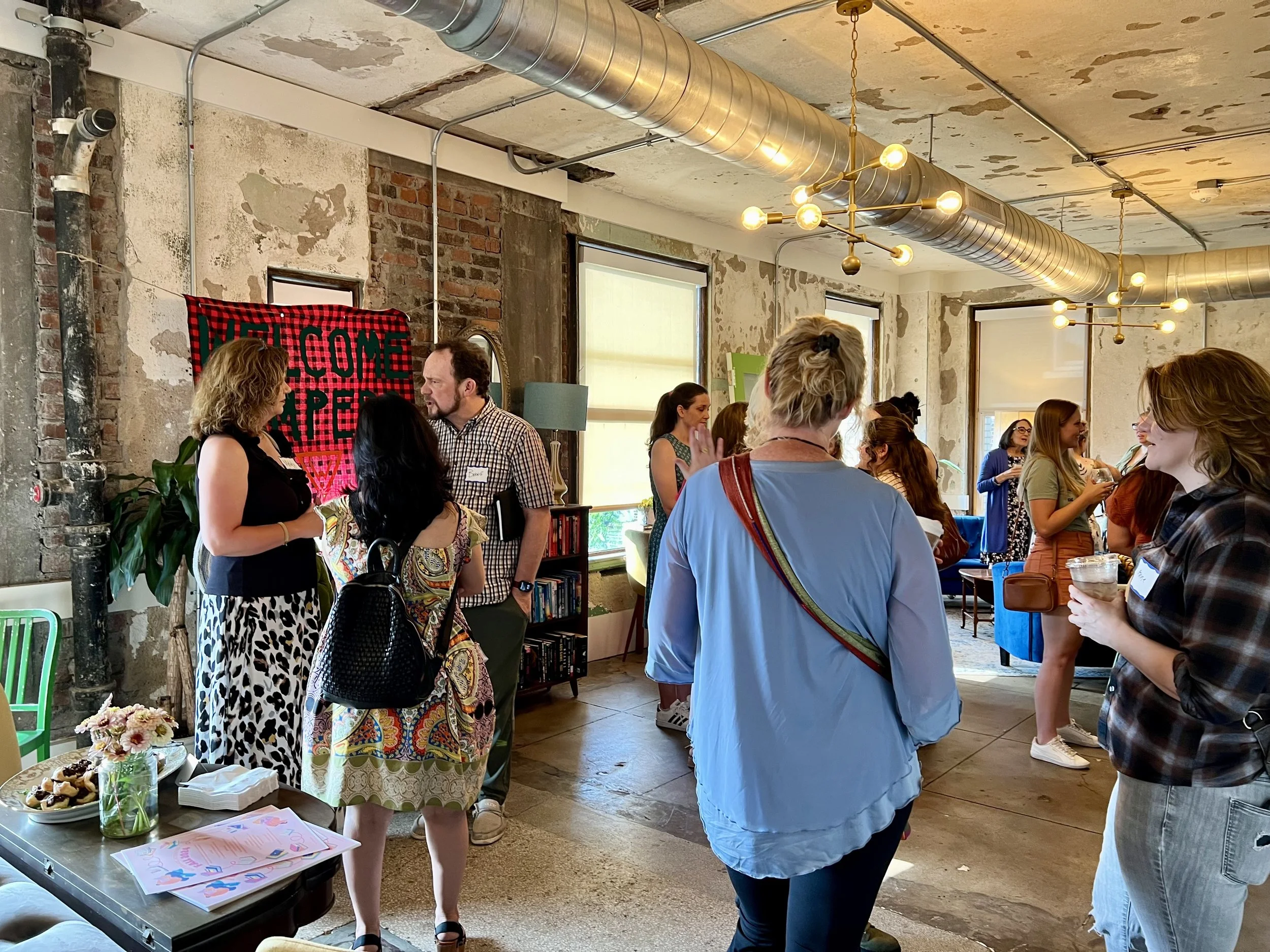 People socializing in an indoor space with exposed brick walls and industrial ceiling, some holding drinks, with a red and black checkered banner that reads 'Welcome PEP' in the background.