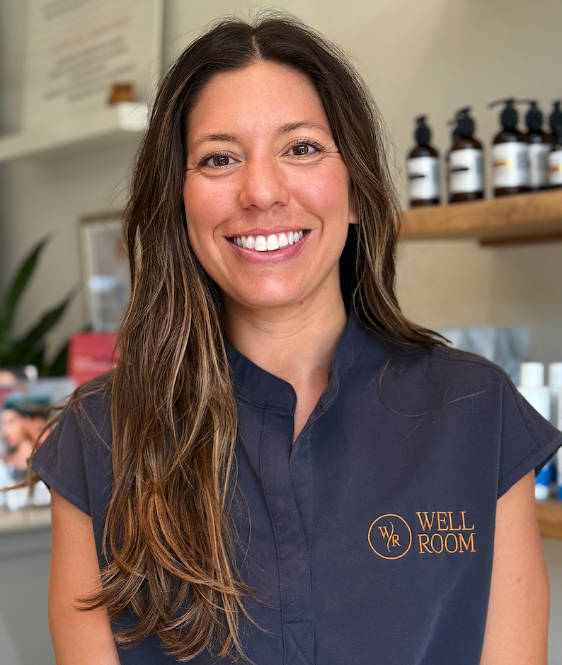 A smiling woman with long brown hair, wearing a dark blue sterile uniform with a "Well Room" logo, standing in a health or wellness clinic.
Well Room IV drip nurse.
