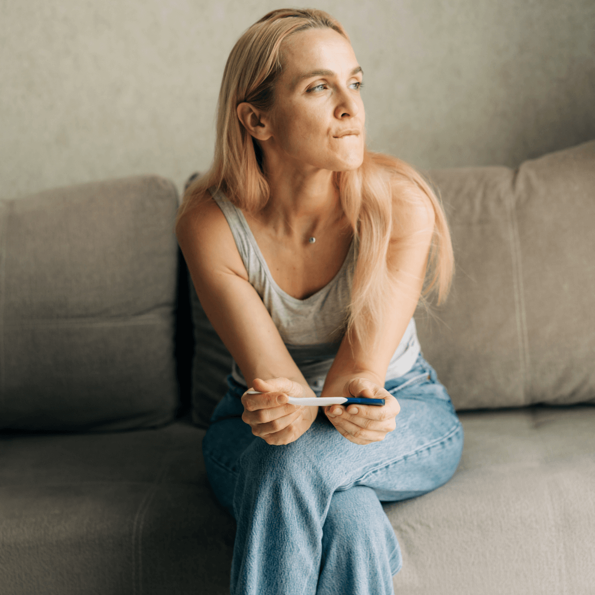 A young woman sitting on a couch with a pregnancy test in her hands thoughtfully looking out to the side