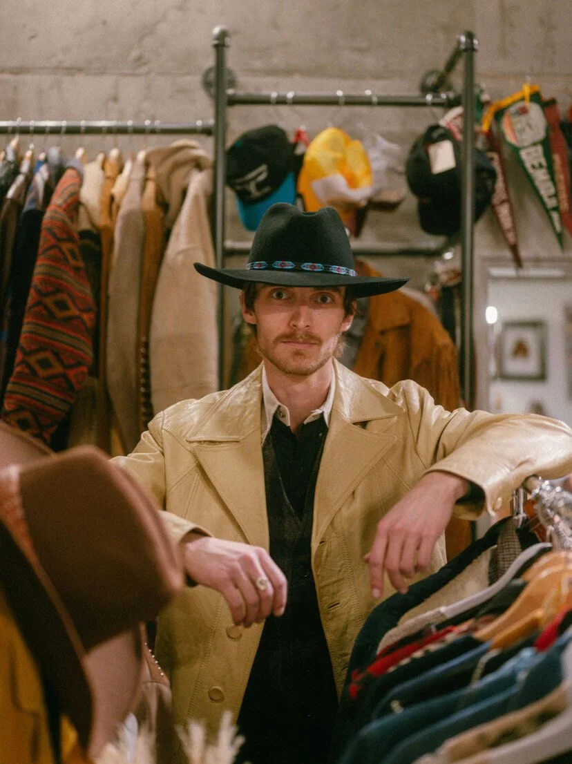 A man wearing a large black hat and beige trench coat shopping in a vintage clothing store.