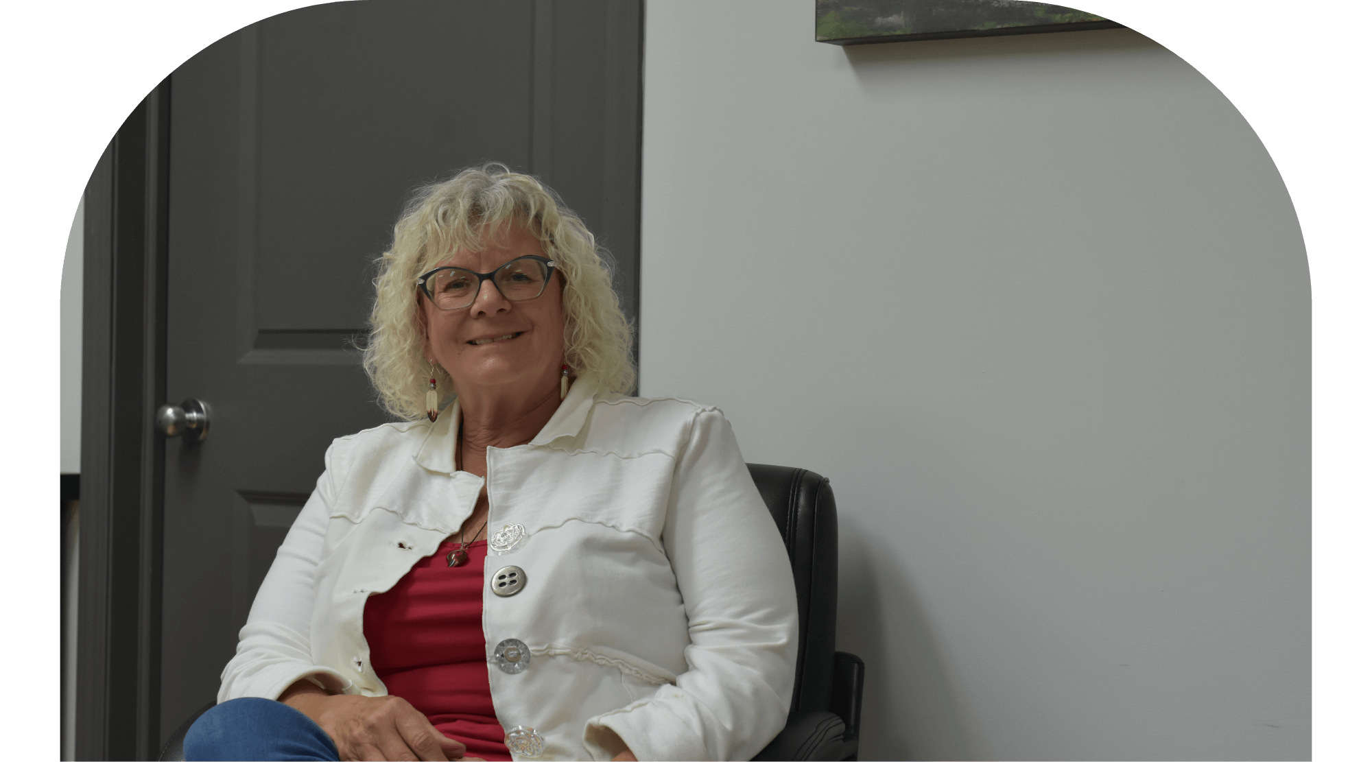 Smiling woman with curly hair wearing glasses and a white jacket sitting in an office chair.