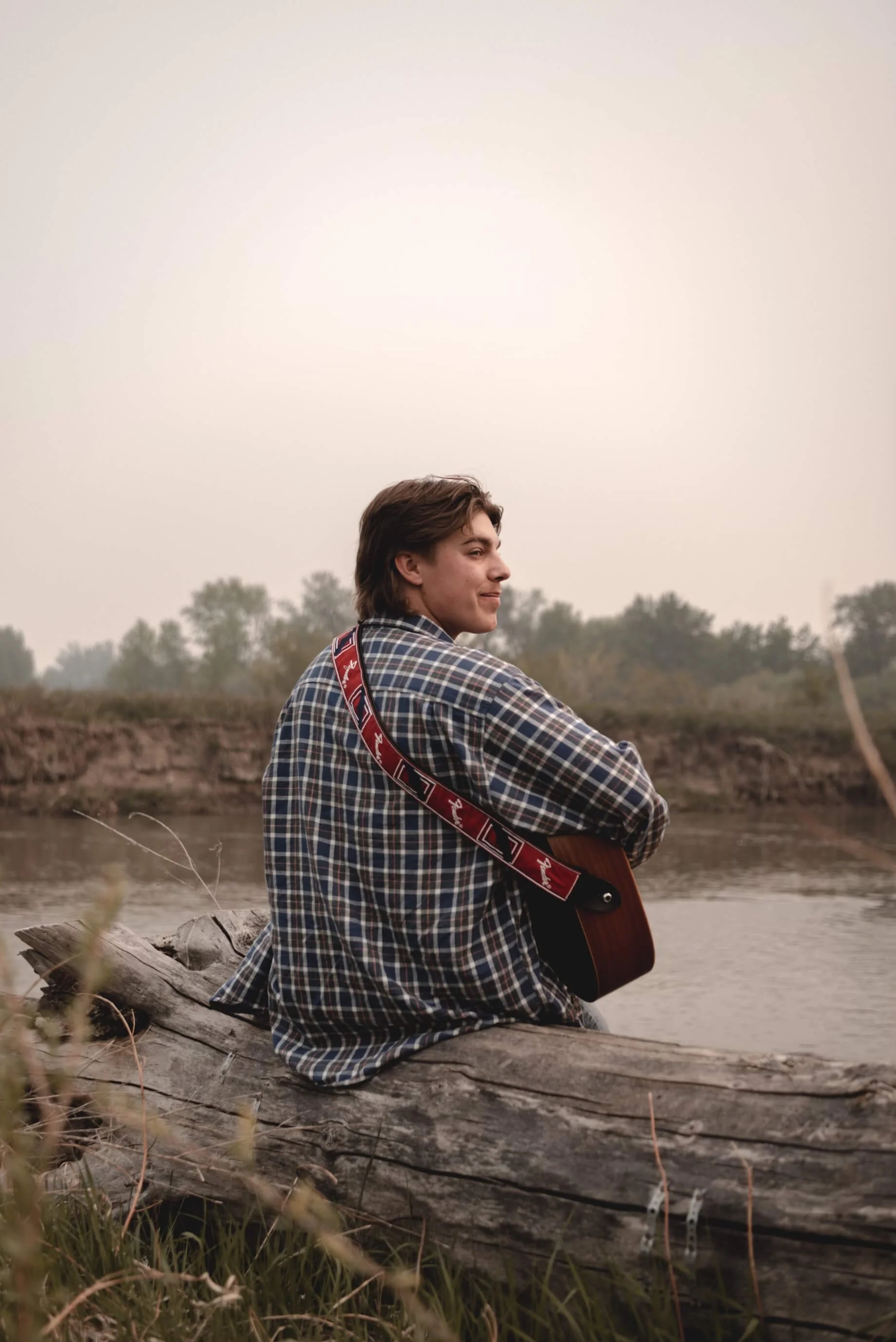 A young man with brown hair sitting on a fallen tree beside a river, holding an acoustic guitar, wearing a checkered shirt, with a cloudy sky and trees in the background.