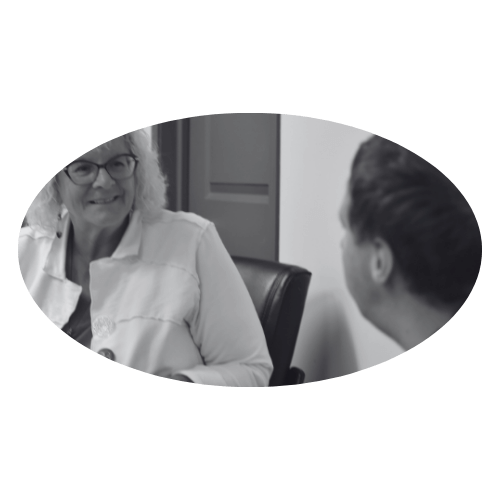 Two people in discussion, one older woman with glasses and curly hair, and a younger person, in an office setting, black and white image.